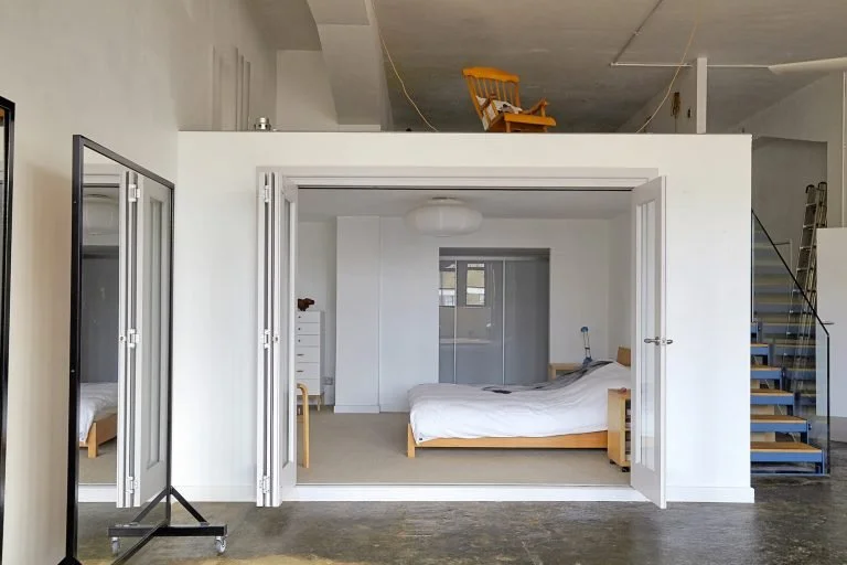 Interior view of a studio apartment with a bed, a white dresser, a mirror, and a staircase leading up to a loft with a yellow chair.