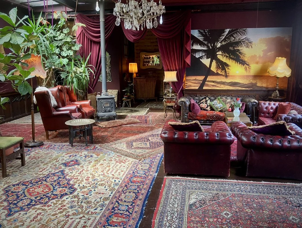 Vintage-style living room with Oriental rugs, leather armchairs and sofas, large potted plants, a chandelier, and a sunset mural of a tropical beach with palm trees.