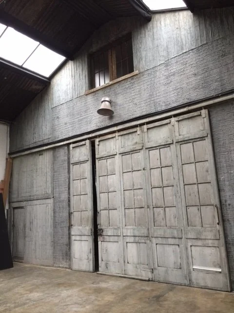 Old wooden barn door with grey paint, in a rustic building with windows and a wall-mounted lamp.