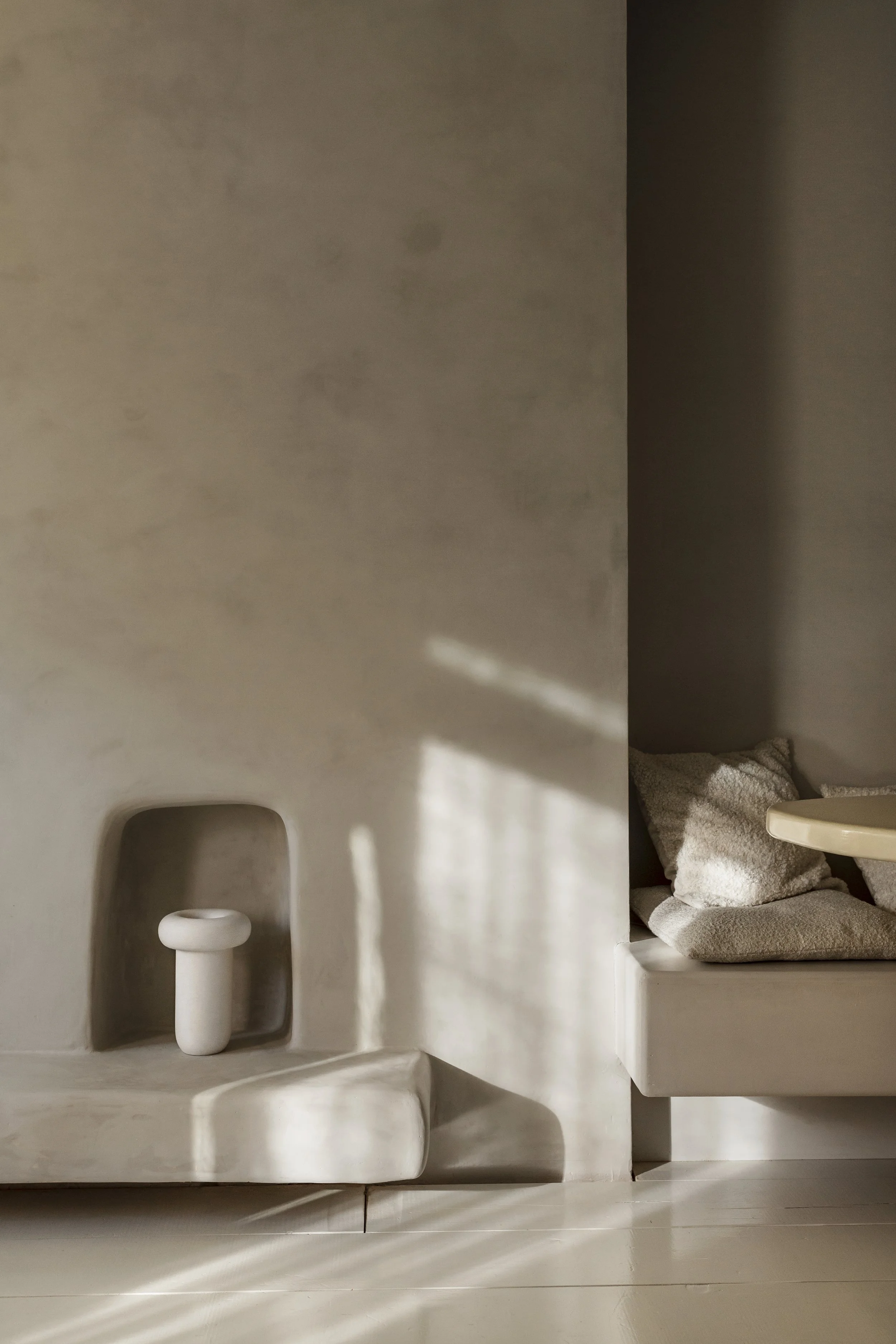 Minimalist interior scene with a built-in niche containing a white ceramic object, possibly a small sculpture or vase, with sunlight casting shadows on a beige wall and corner seating area with pillows.