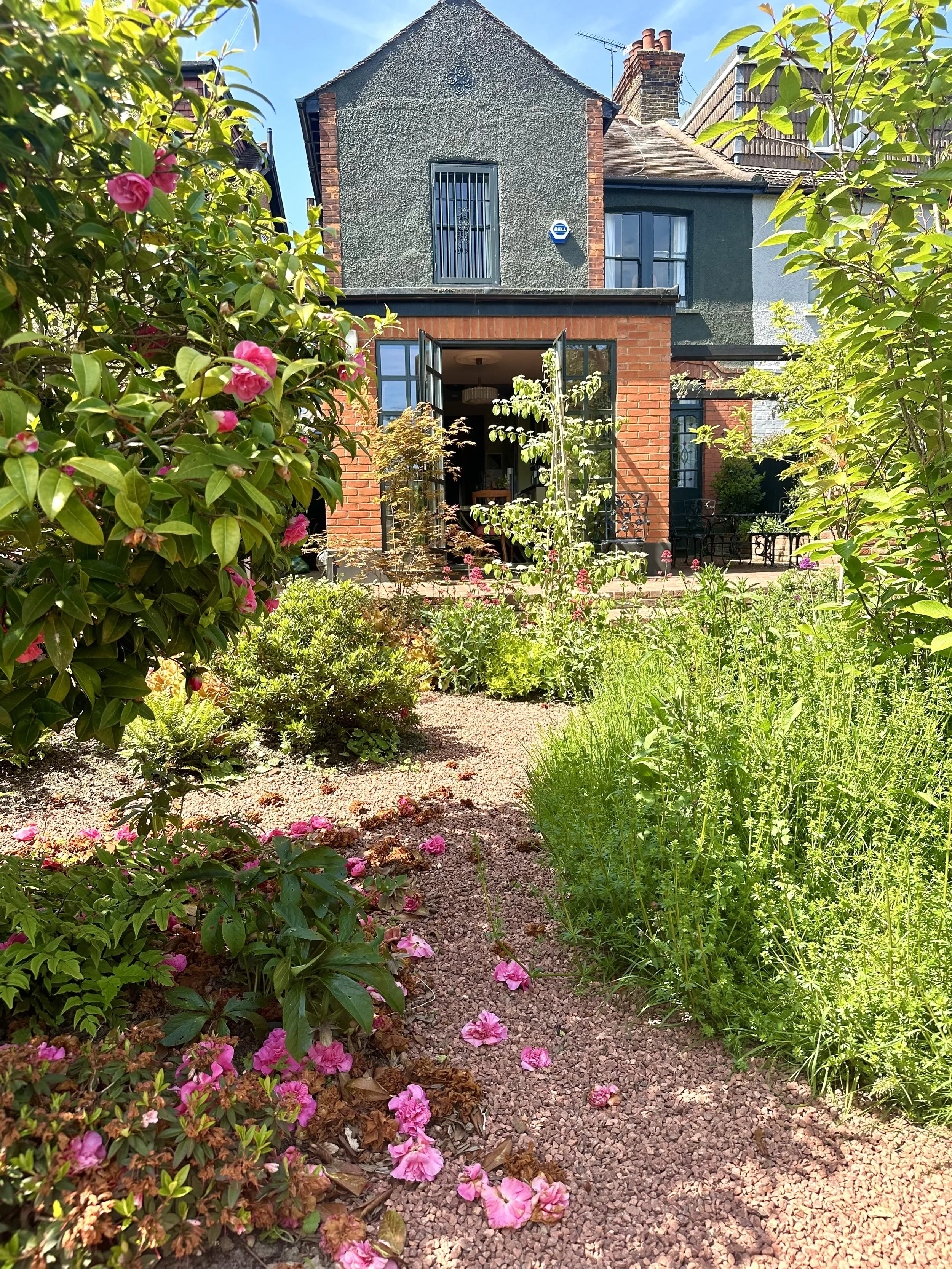 A view of a garden pathway leading up to a house with a brick and dark grey exterior, surrounded by green plants and pink flowers, under a clear blue sky.