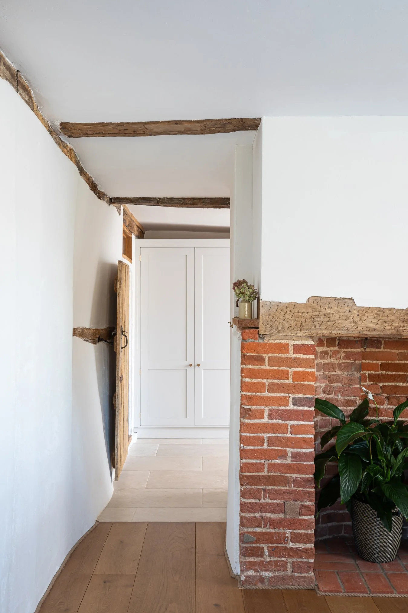 Interior view of a room with white walls, wooden beams on the ceiling, a white cabinet, and a brick fireplace with a potted plant nearby.