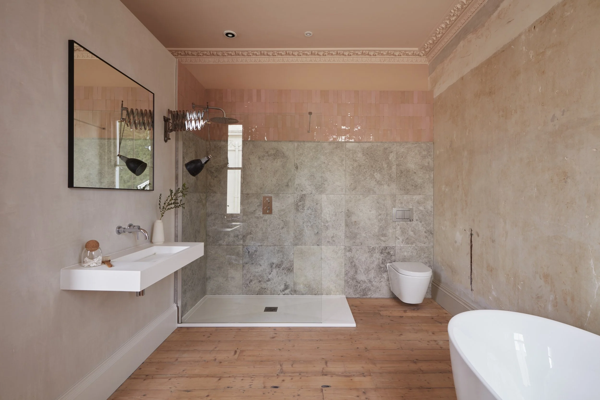 Minimalist bathroom with wooden floor, wall-mounted white sink, large mirror, black wall lamps, walk-in shower with gray tiles, wall-mounted toilet, and a freestanding bathtub. Pink and beige walls with ornate crown molding.