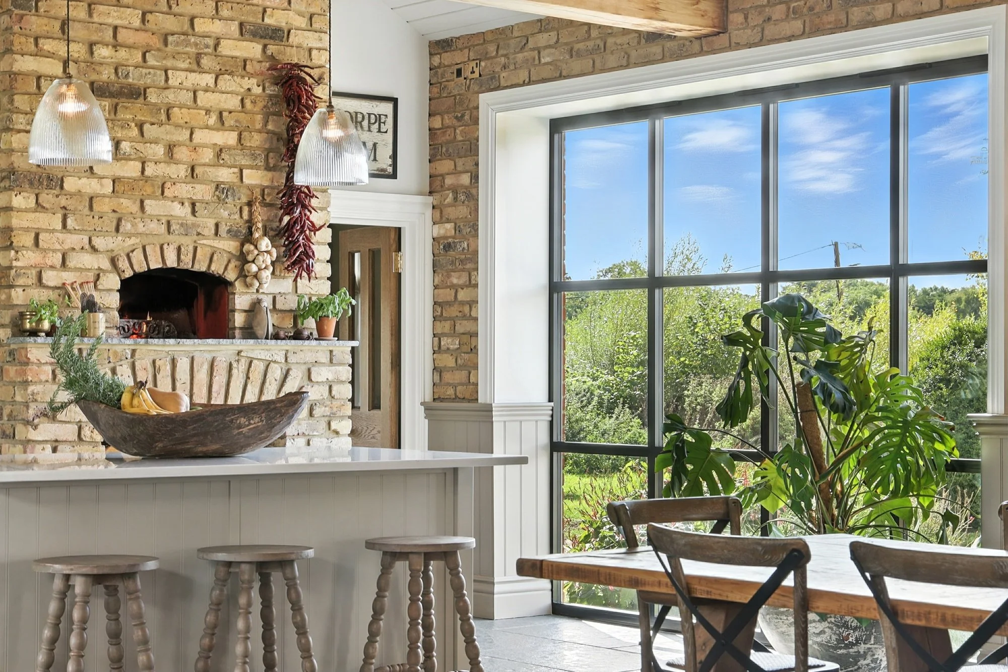 A cozy kitchen with a brick wall and large window overlooking a green outdoor area. The counter has a wooden bowl with bananas and herbs, and various kitchen items.