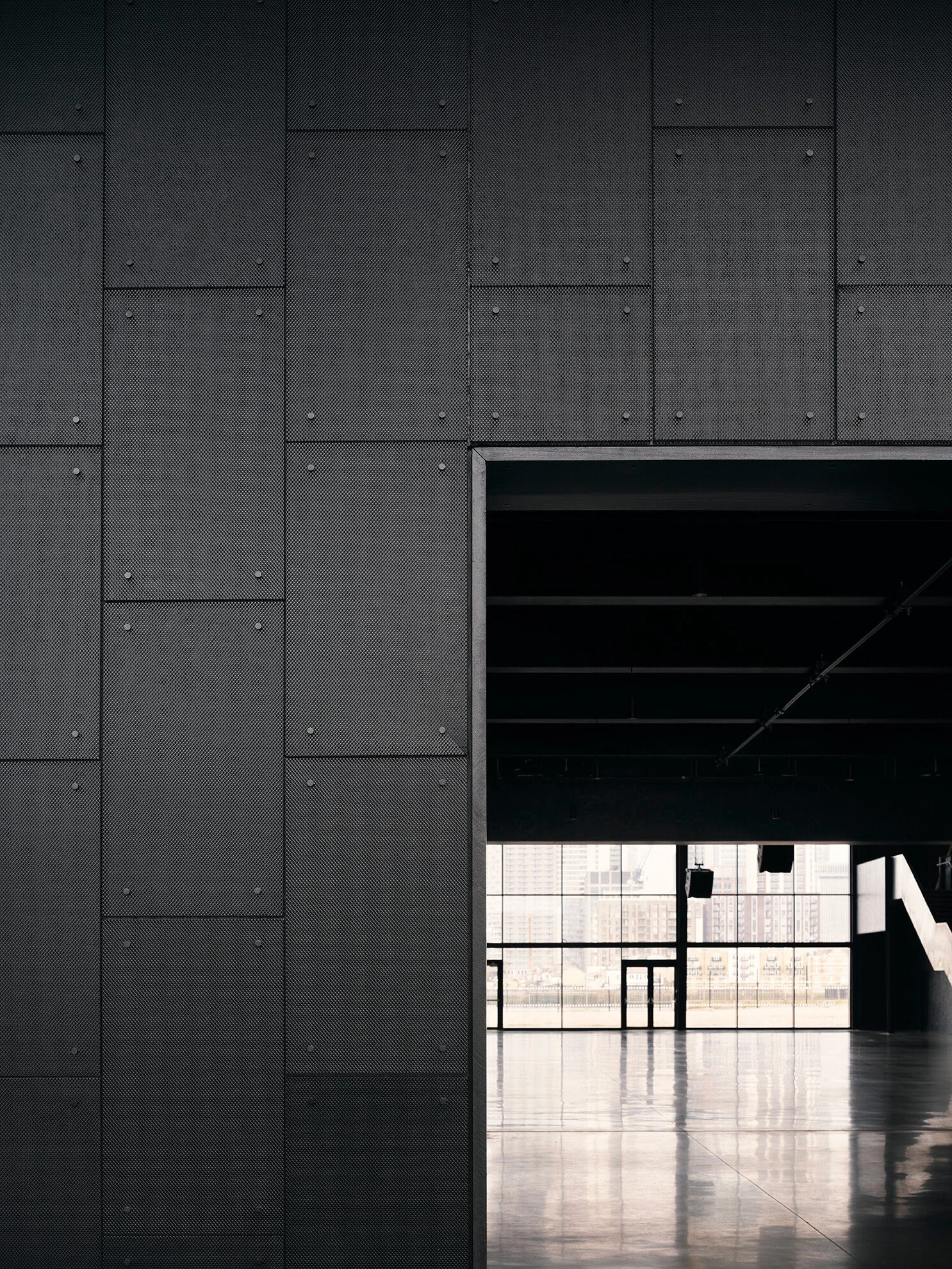 Interior view of a modern building with black textured walls, a large open doorway, and windows showing a cityscape outside.