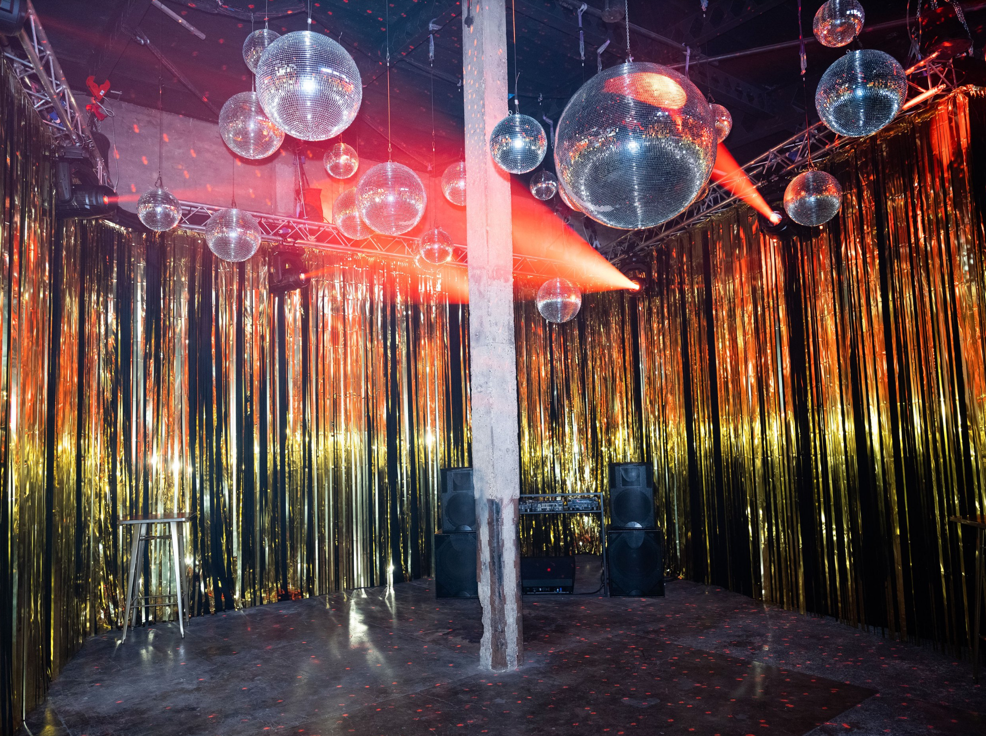 An empty nightclub or party space decorated with shiny gold and black streamers and several disco balls hanging from the ceiling, with colorful lighting effects.