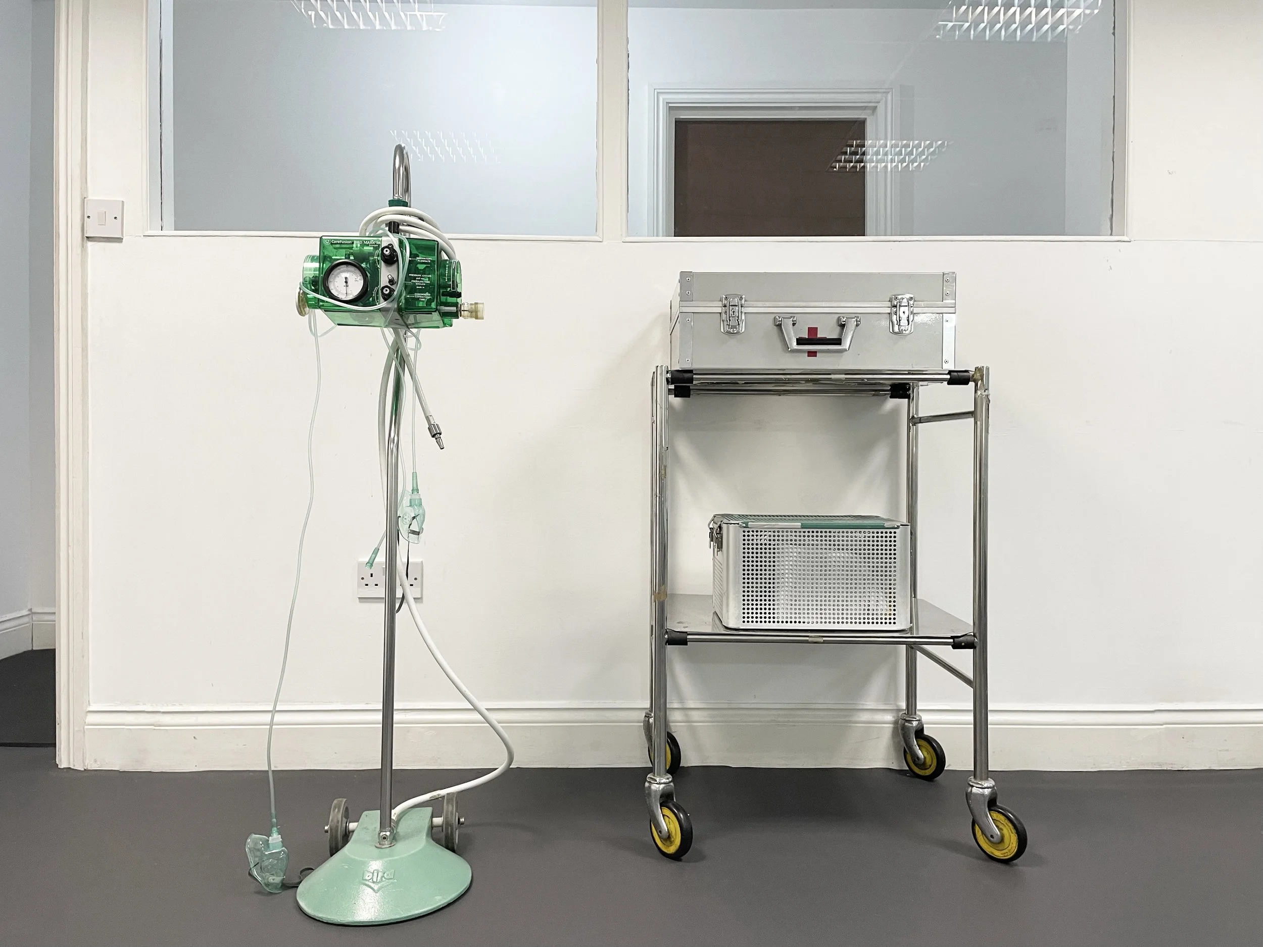 Medical IV stand with green infusion pump and a hospital trolley with a metal box and a lower shelf, on a gray floor against a white wall with windows.