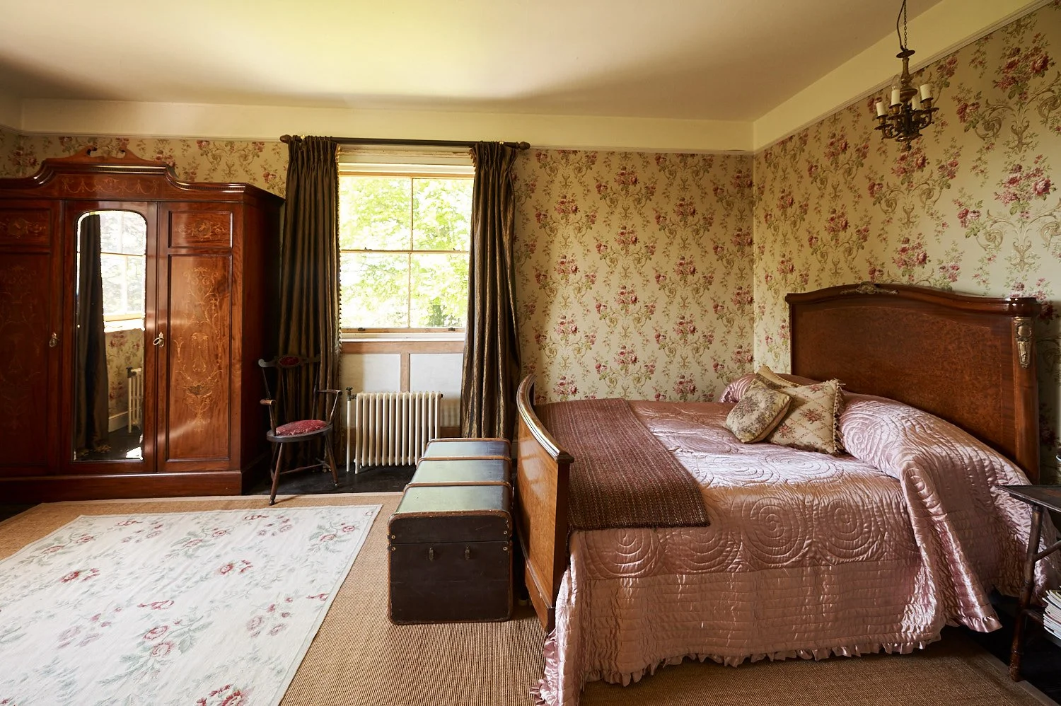 Vintage bedroom with floral wallpaper, wooden bed with satin bedding, armoire, window with curtains, vintage chair, radiator, and trunk at the foot of the bed.