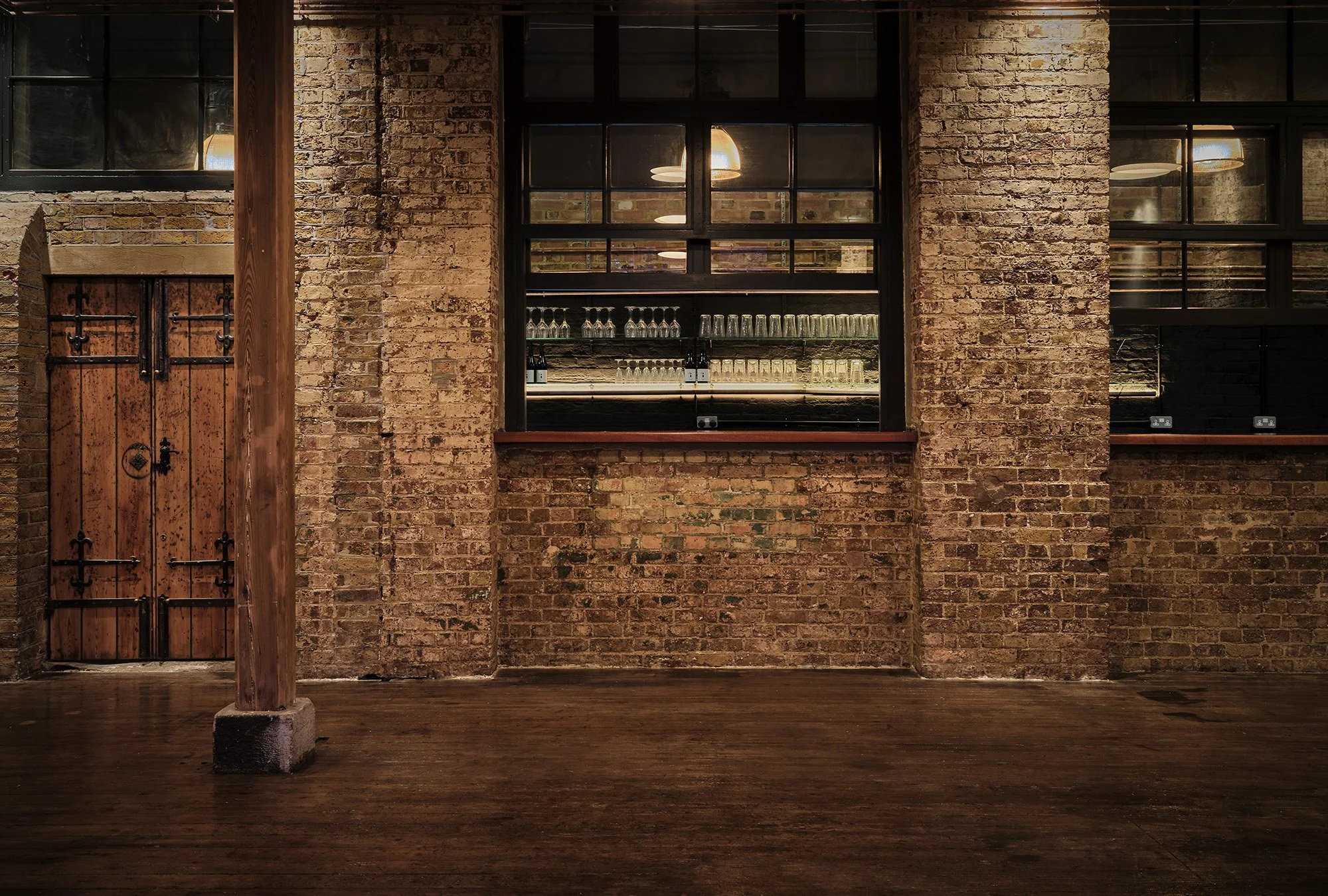 Interior of a brick-walled industrial-style space with a dark wooden floor, a wooden door on the left, a large window in the center showing glasses and bottles, and exposed brick walls.