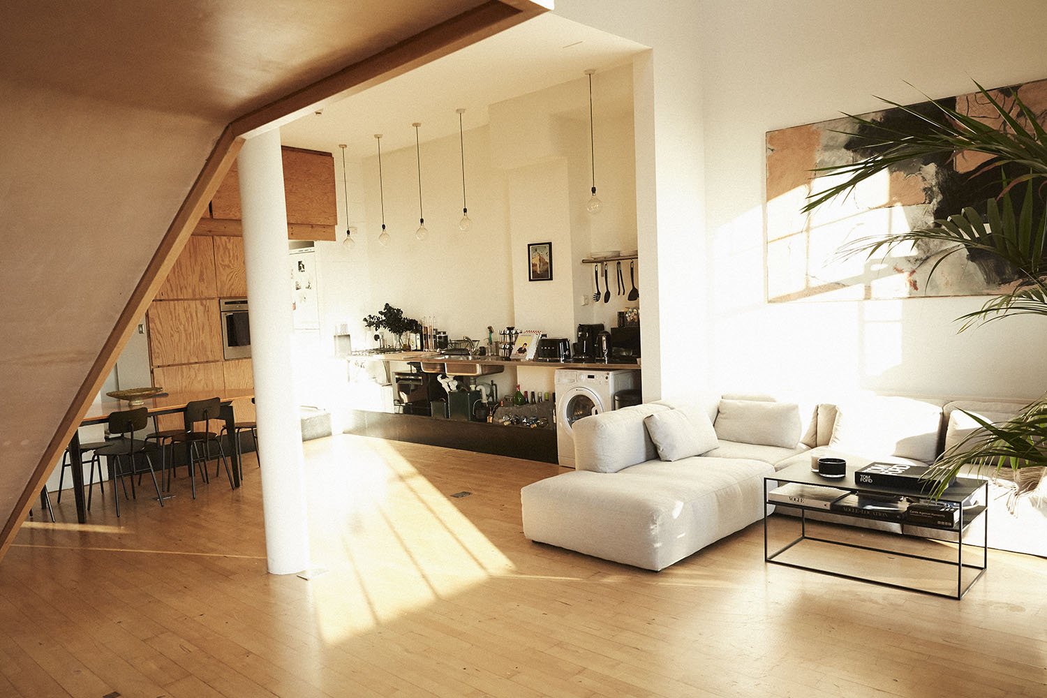Living room with a white sectional sofa near a large window, wooden flooring, and a modern open kitchen in the background with hanging pendant lights, kitchen appliances, and a dining area with chairs.