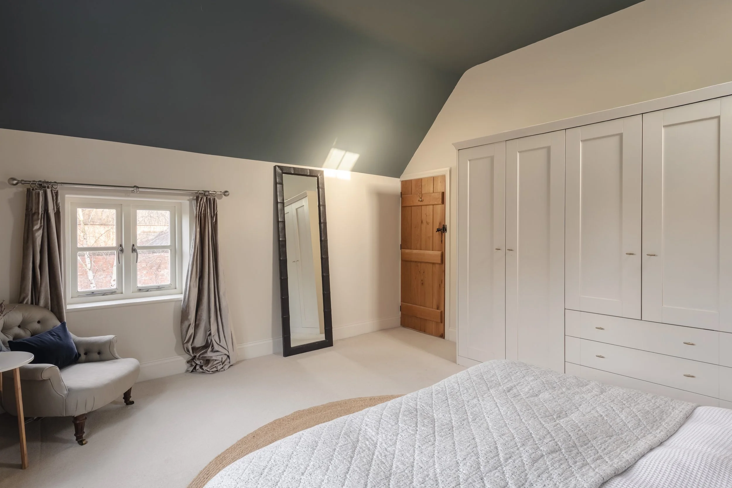 A bedroom with a sloped ceiling, a large white wardrobe with multiple doors and drawers, a small window with curtains, a beige armchair with a dark pillow, a floor-standing mirror, and a wooden door.