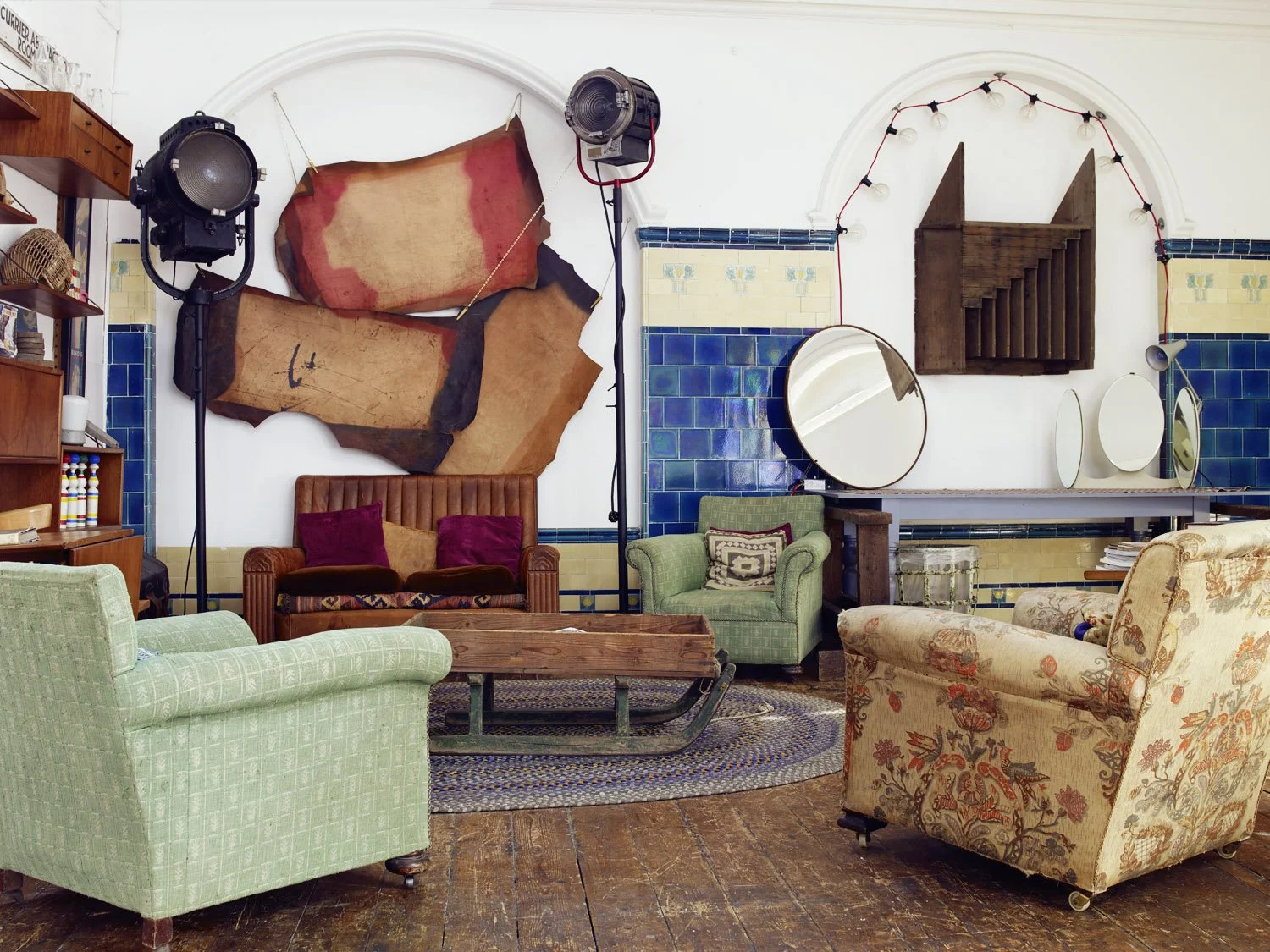Eclectic living room with vintage furniture, including a green armchair, floral armchair, and brown leather sofa, a wooden coffee table, and decorative mirrors on the wall. Large stage lights are positioned on stands, and a rustic wooden wall art pie
