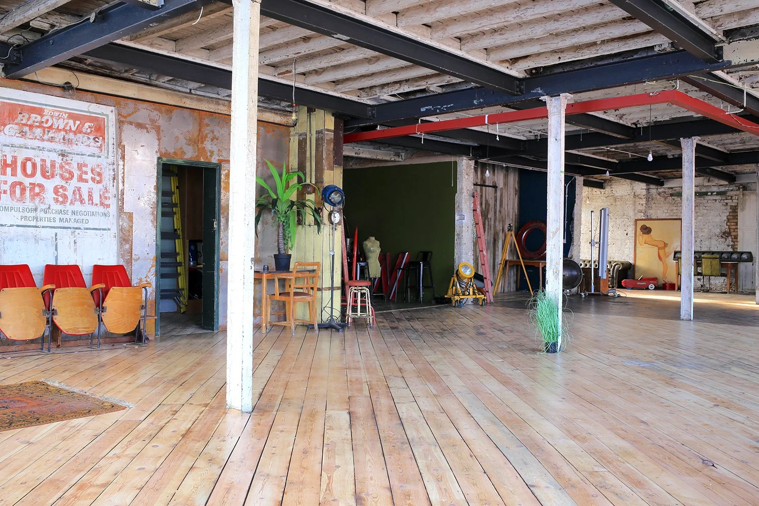 Interior of an industrial-style space with wooden floors, exposed beams, and eclectic furniture, including red seats, chairs, plants, and gym equipment in the background.