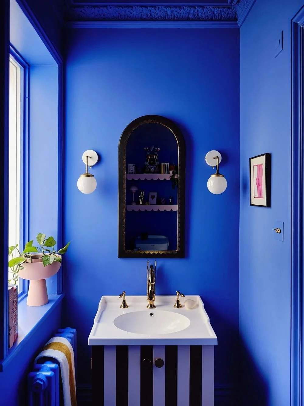 Blue bathroom with a window, a white sink with gold fixtures, striped black and white cabinet, a mirror, two white wall sconces, a framed pink artwork, a pink pot with a green plant, and a radiator with a yellow and white towel.