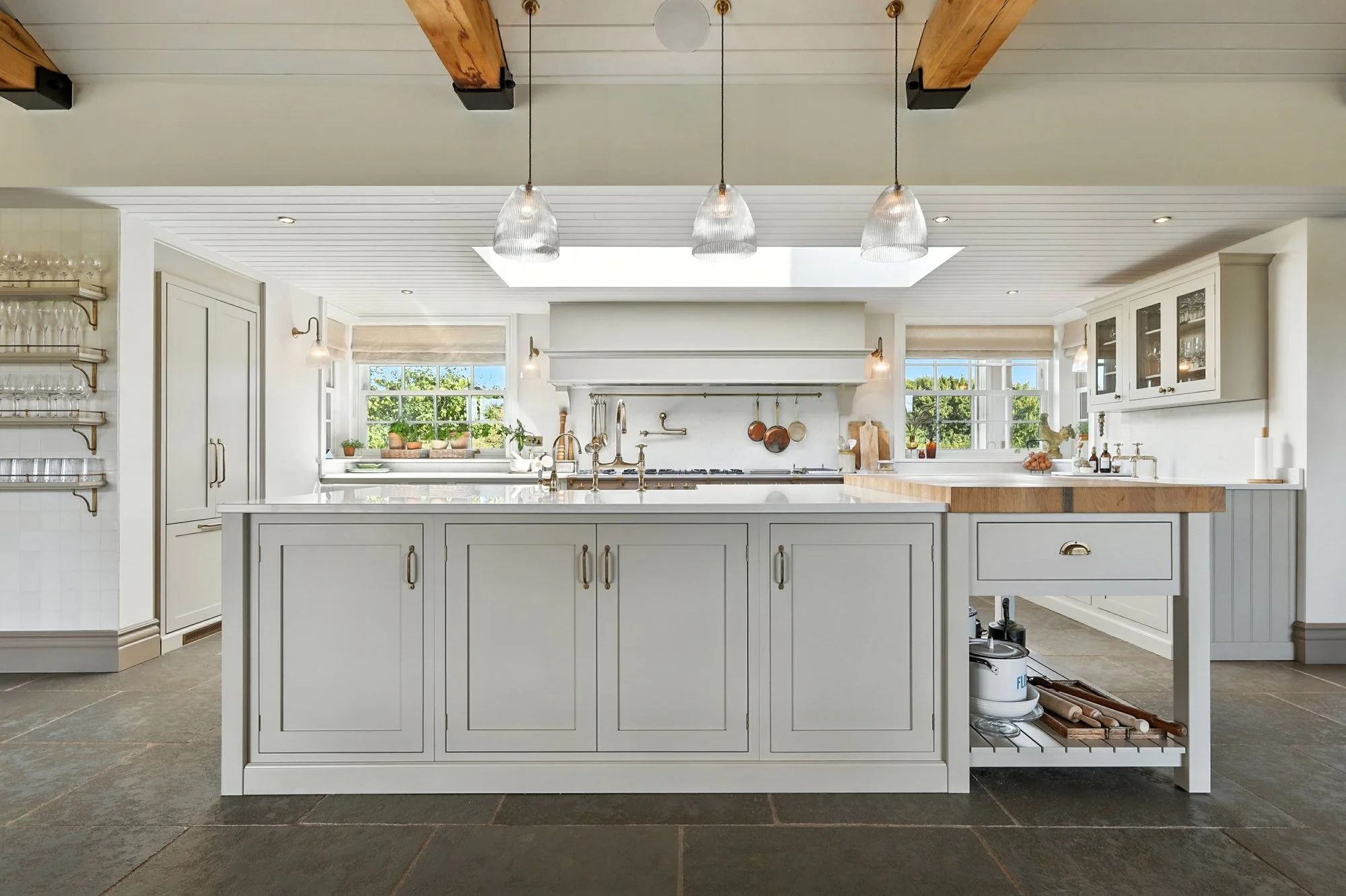 A bright kitchen with white cabinets, a large island, and three pendant lights hanging from a wooden-beamed ceiling. There are windows with blinds letting in natural light, and kitchen utensils hanging on the wall.