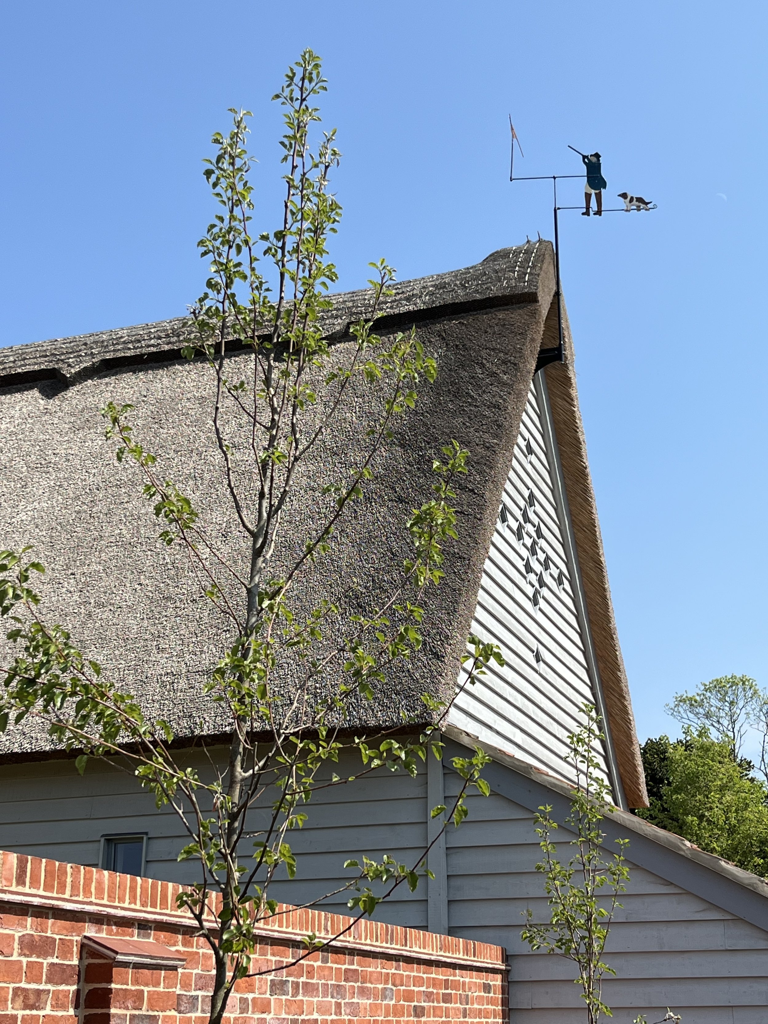 A house with a thatched roof and a weather vane on top featuring a person fishing with a dog, set against a clear blue sky. There are trees and a brick wall in the foreground.