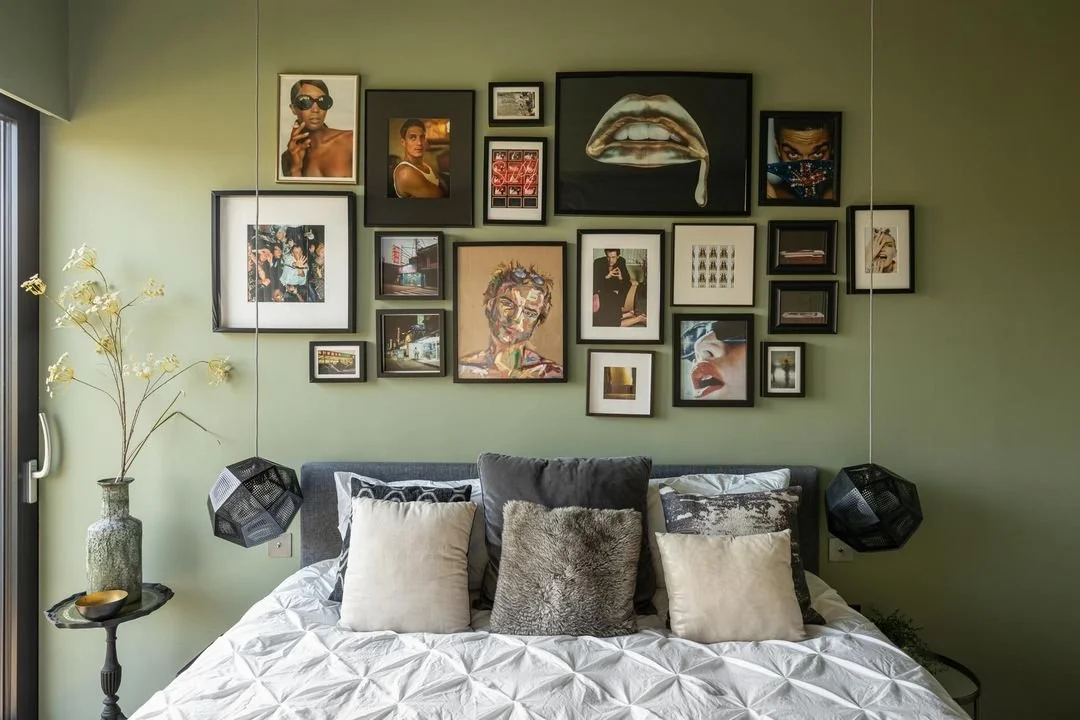 A bedroom with a green wall decorated with a gallery of framed photos and artwork, including portraits, abstract art, and illustrations. A bed with white bedding and multiple pillows sits below the gallery, with black pendant lights hanging on either