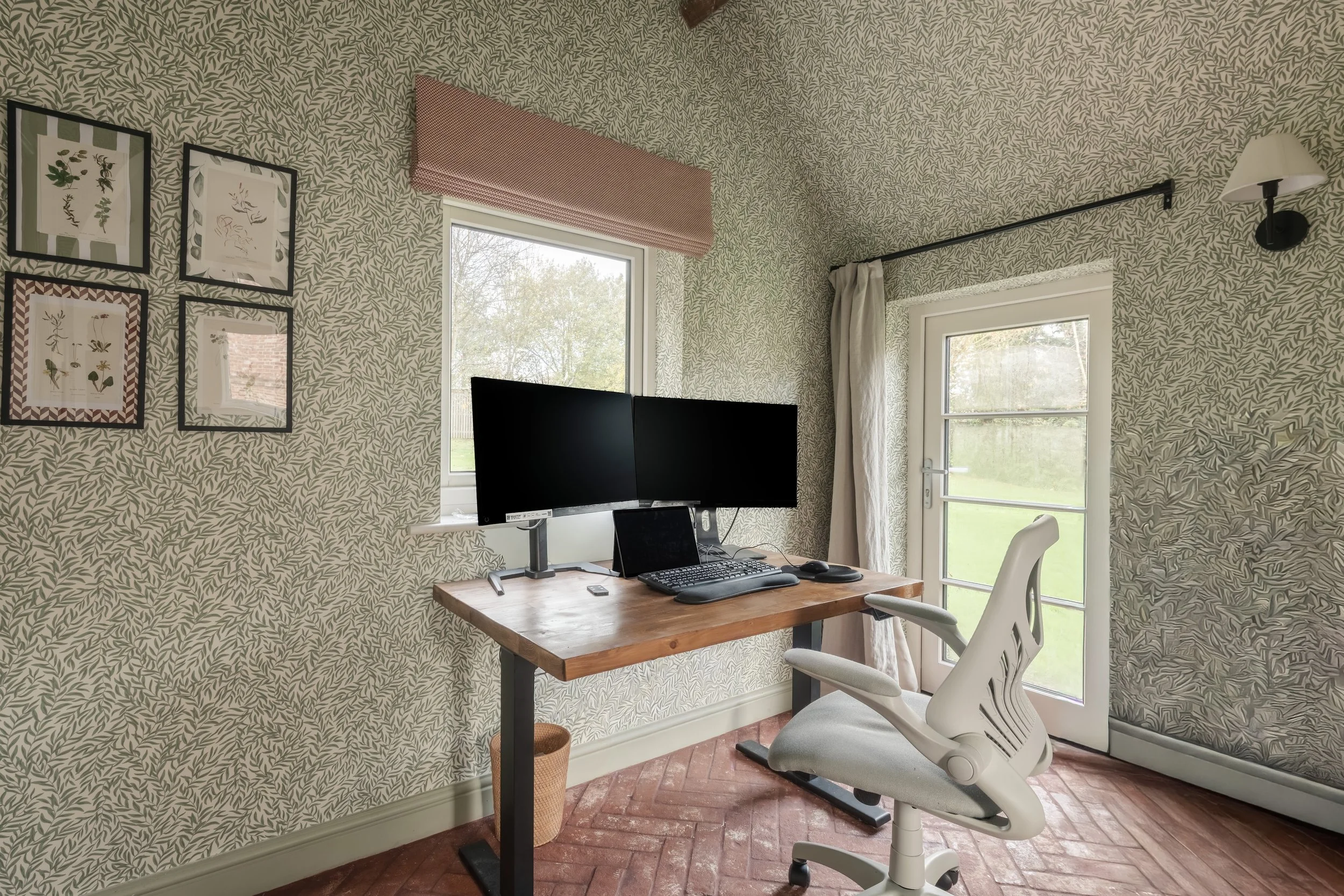 Home office with a wooden desk, dual monitors, ergonomic office chair, and wallpaper with a leaf pattern; window and door with curtains, wall-mounted lamp, and framed botanical art