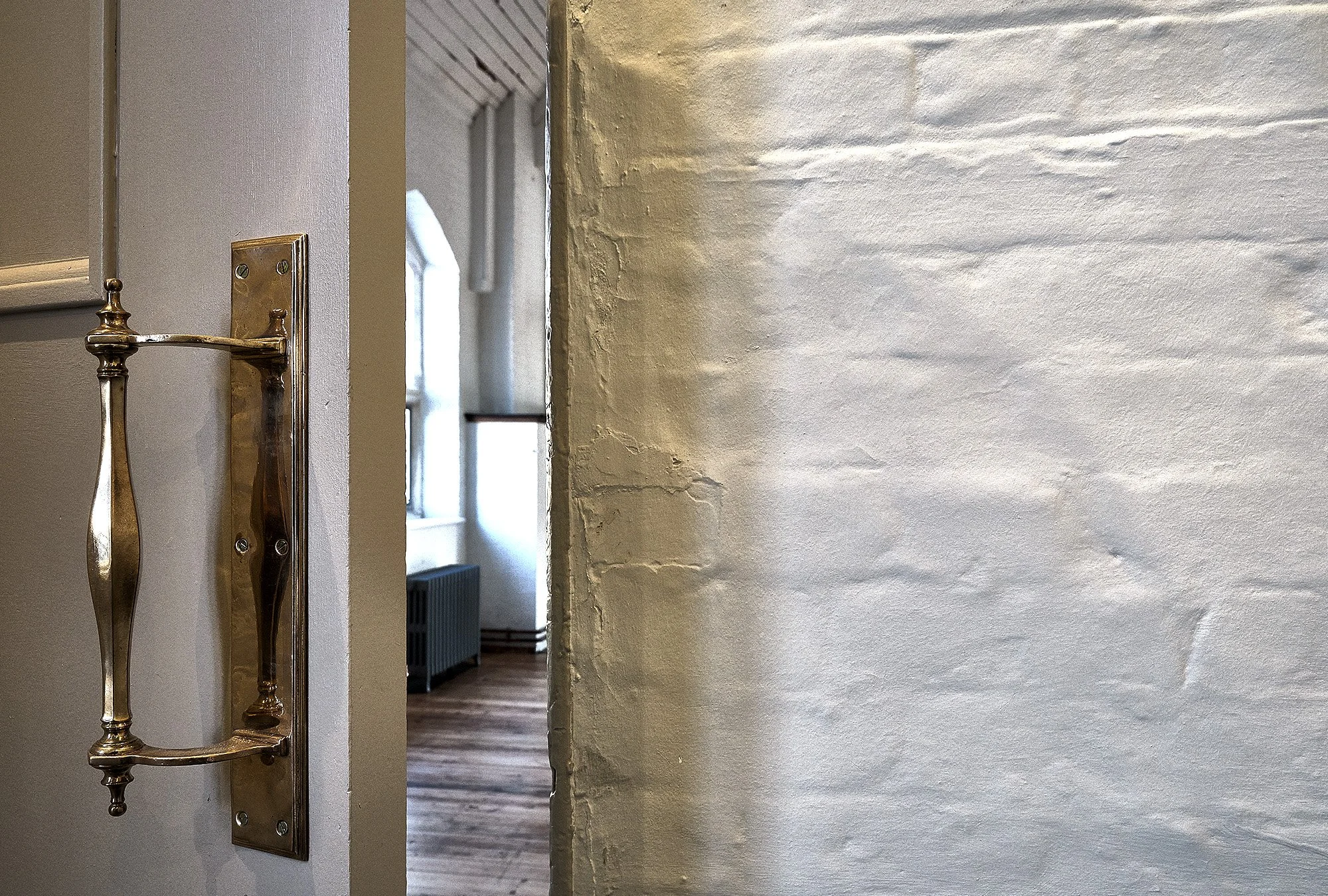 Interior view showing a metallic towel rack mounted on a white wall, a doorway revealing a room with a wooden floor, a window, and a radiator, with a textured white brick wall on the right.