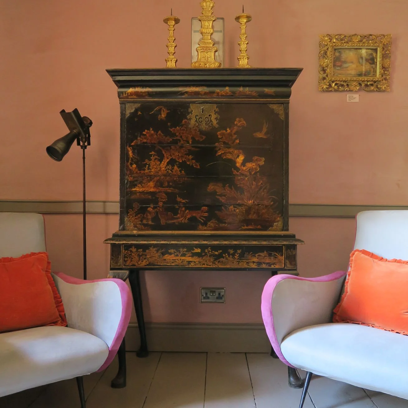 Decorative black lacquered cabinet with intricate gold landscape painting on it, flanked by two white chairs with pink and orange cushions, against a pink wall with framed artwork and a small casting lamp on a stand.
