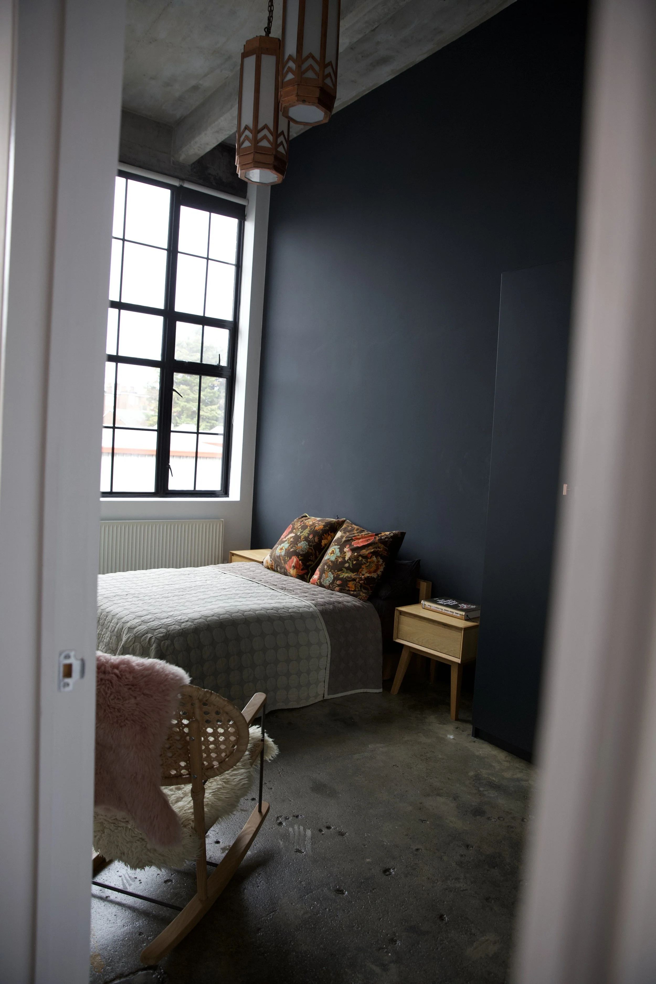 A bedroom with a dark accent wall, large window, bed with floral pillows, wooden nightstands, a rocking chair with a pink fluffy throw, and pendant lights hanging from the ceiling.