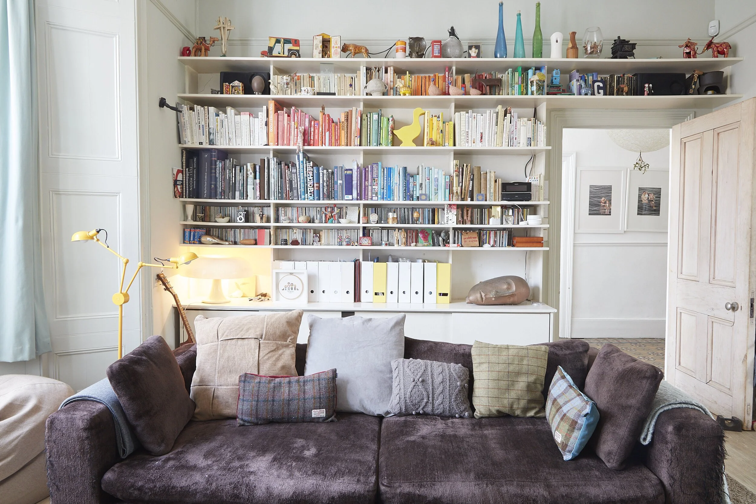 A cozy living room scene featuring a dark brown plush sofa with multiple pillows, a yellow adjustable floor lamp, a white side table with a lamp, and a large white bookshelf filled with books, decorative objects, and colorful items. There are also wo