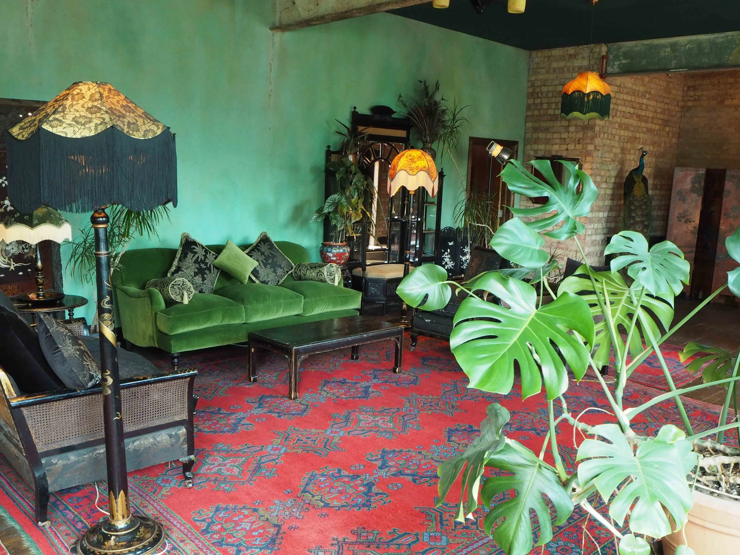 A living room with green velvet sofa, patterned cushions, large leafy plant, vintage lamps with fringed shades, and decorative elements including a red patterned rug, plants, and antique furniture against brick and green walls.