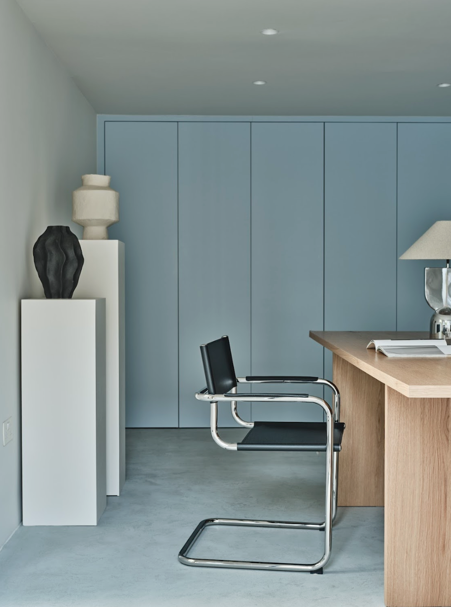 Modern office room with a black desk chair, a wooden desk, blue cabinets, white and black decorative vases, and a table lamp.