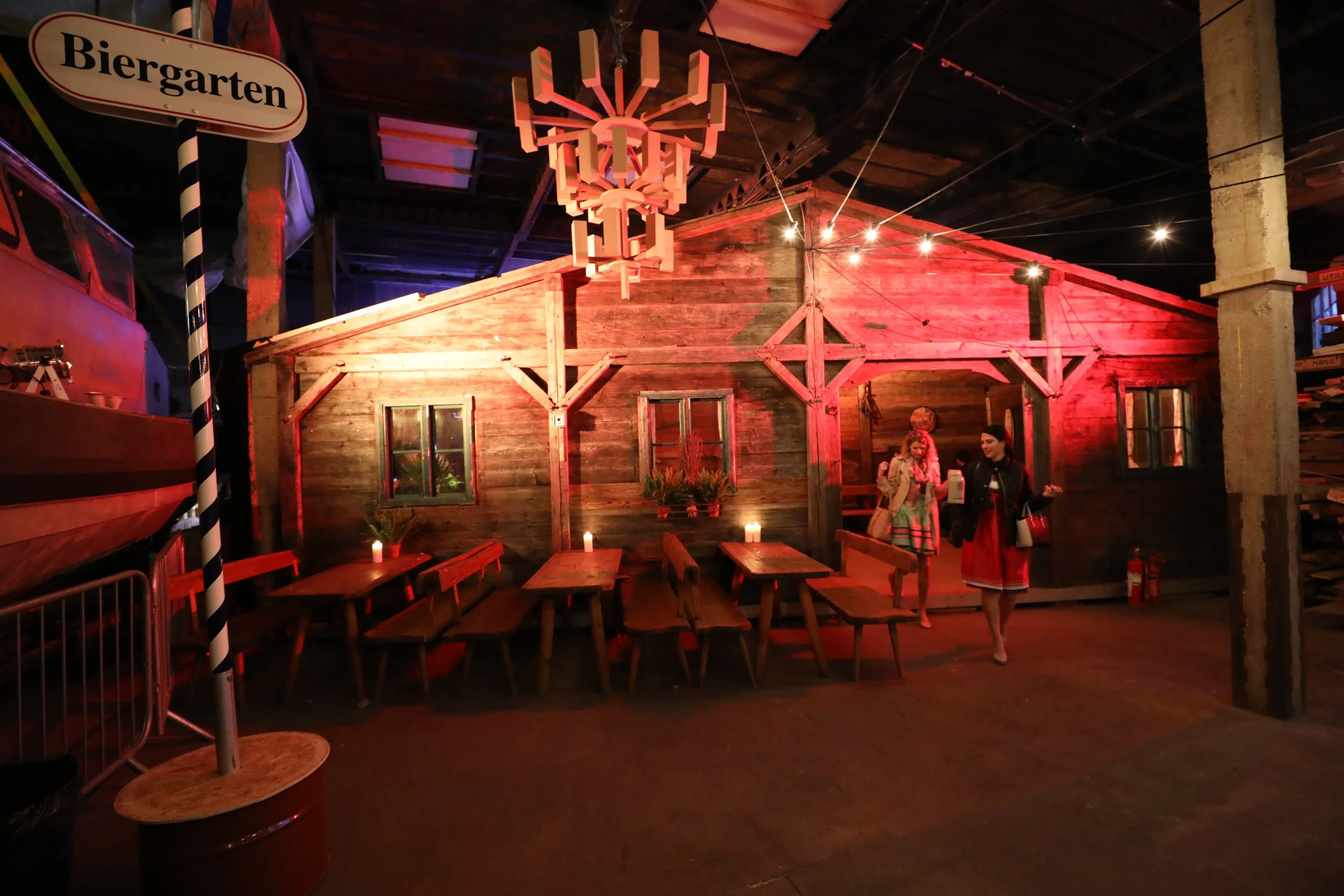 Indoor scene with a rustic wooden hut, a sign reading 'Biergarten', several wooden tables with lit candles, and two women in traditional Bavarian dresses walking inside, illuminated with red and purple lighting.