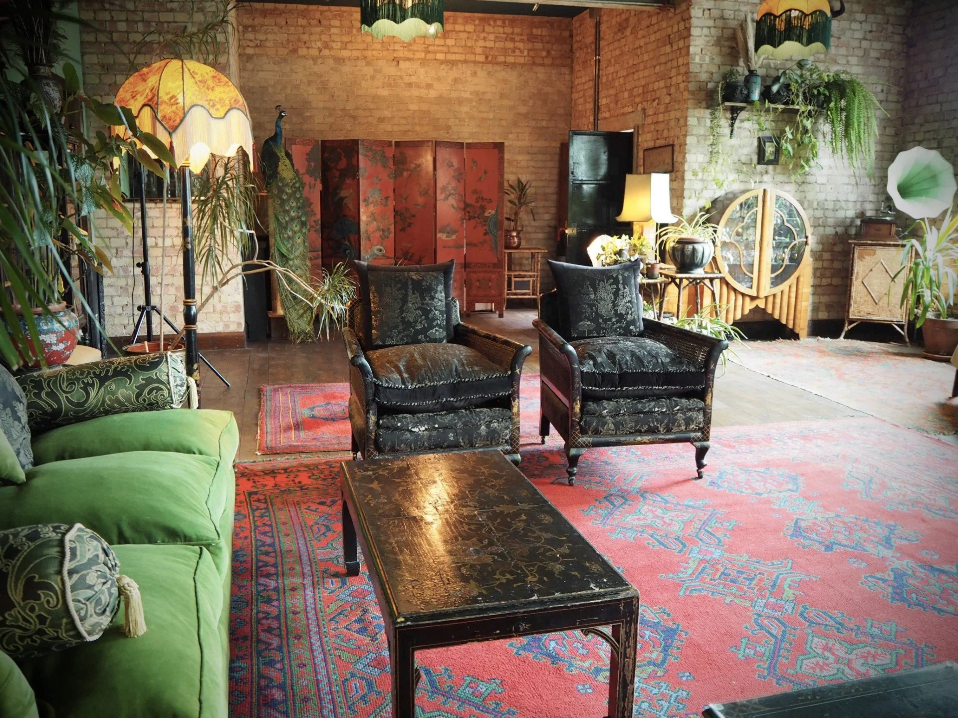 A living room with vintage furniture, including a green sofa, black upholstered armchairs, a black lacquered table, and wooden sideboards. The room has exposed brick walls, hardwood floors, large potted plants, and decorative lamps with shades. There