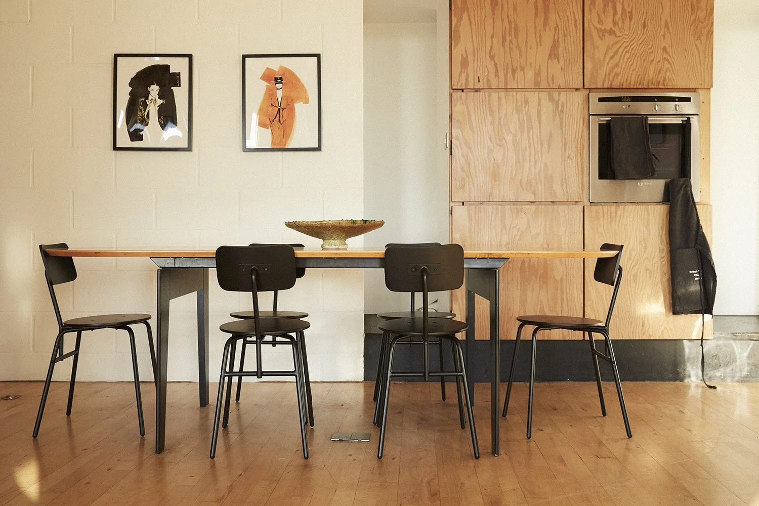 Interior of a dining area in a modern home with a wooden table, five black chairs, a decorative bowl on the table, and wall art featuring stylized portraits. In the background, a wooden cabinet with a built-in oven and a black apron hanging on the si