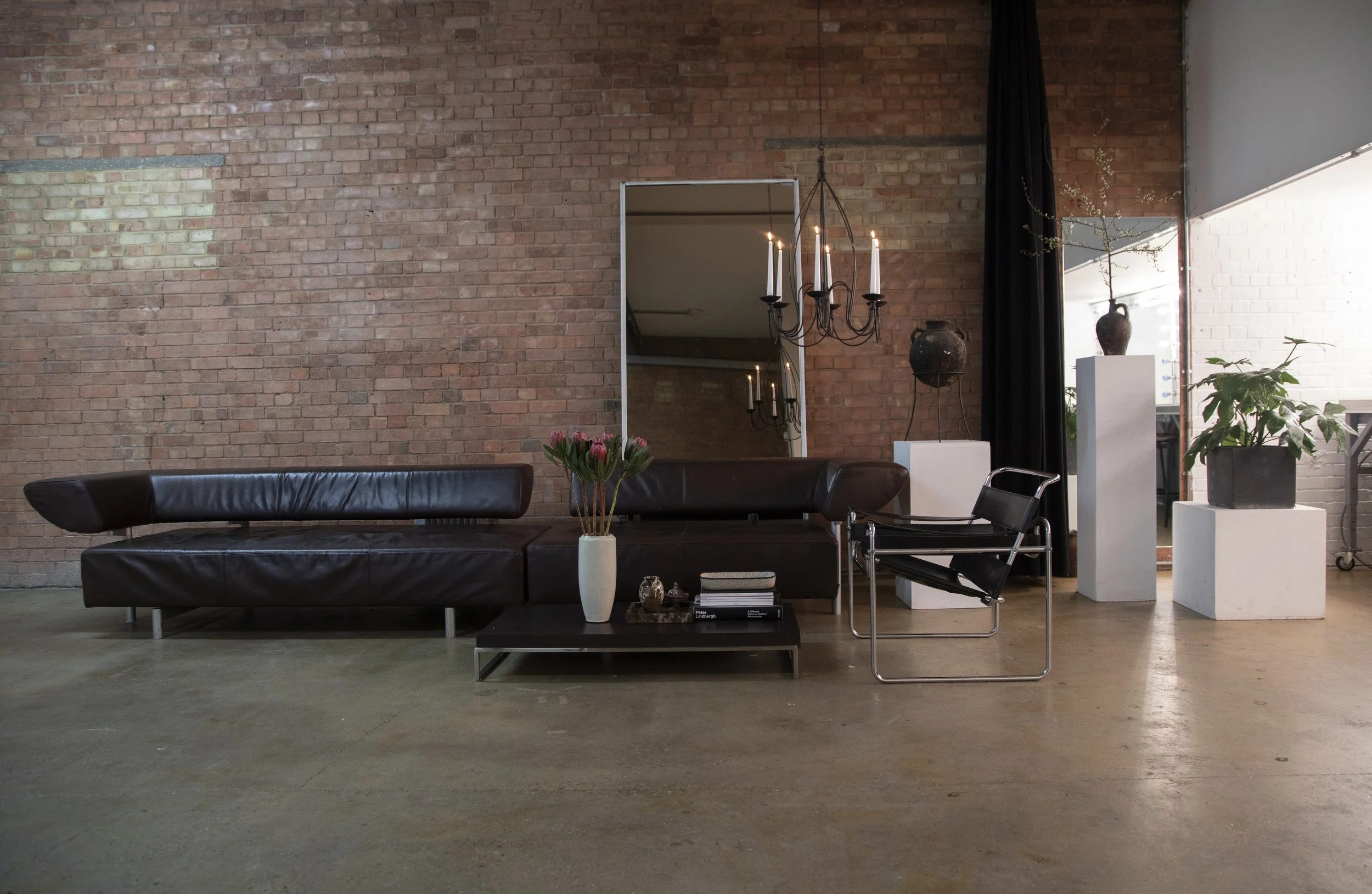 Living room with brick wall, black leather sectional sofa, black coffee table with books and flowers, modern black chair, tall white pedestals with potted plants, large mirror, hanging chandelier with candles, and a large window with natural light.