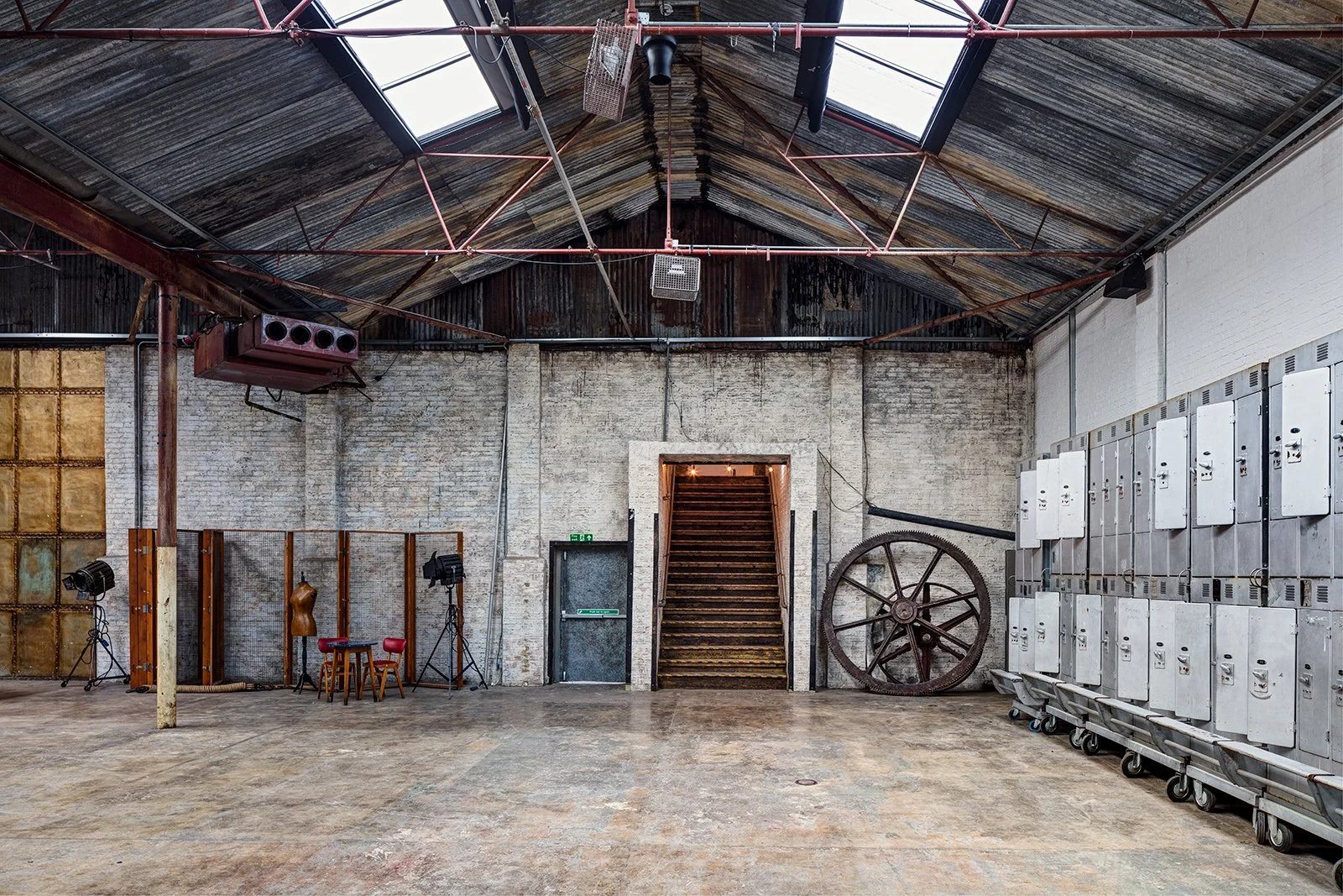 An industrial-style room with a sloped metal roof and brick walls. There are lockers on the right, some lighting equipment, a large wheel leaning against the wall, and a small set of stairs leading up to a doorway at the back.