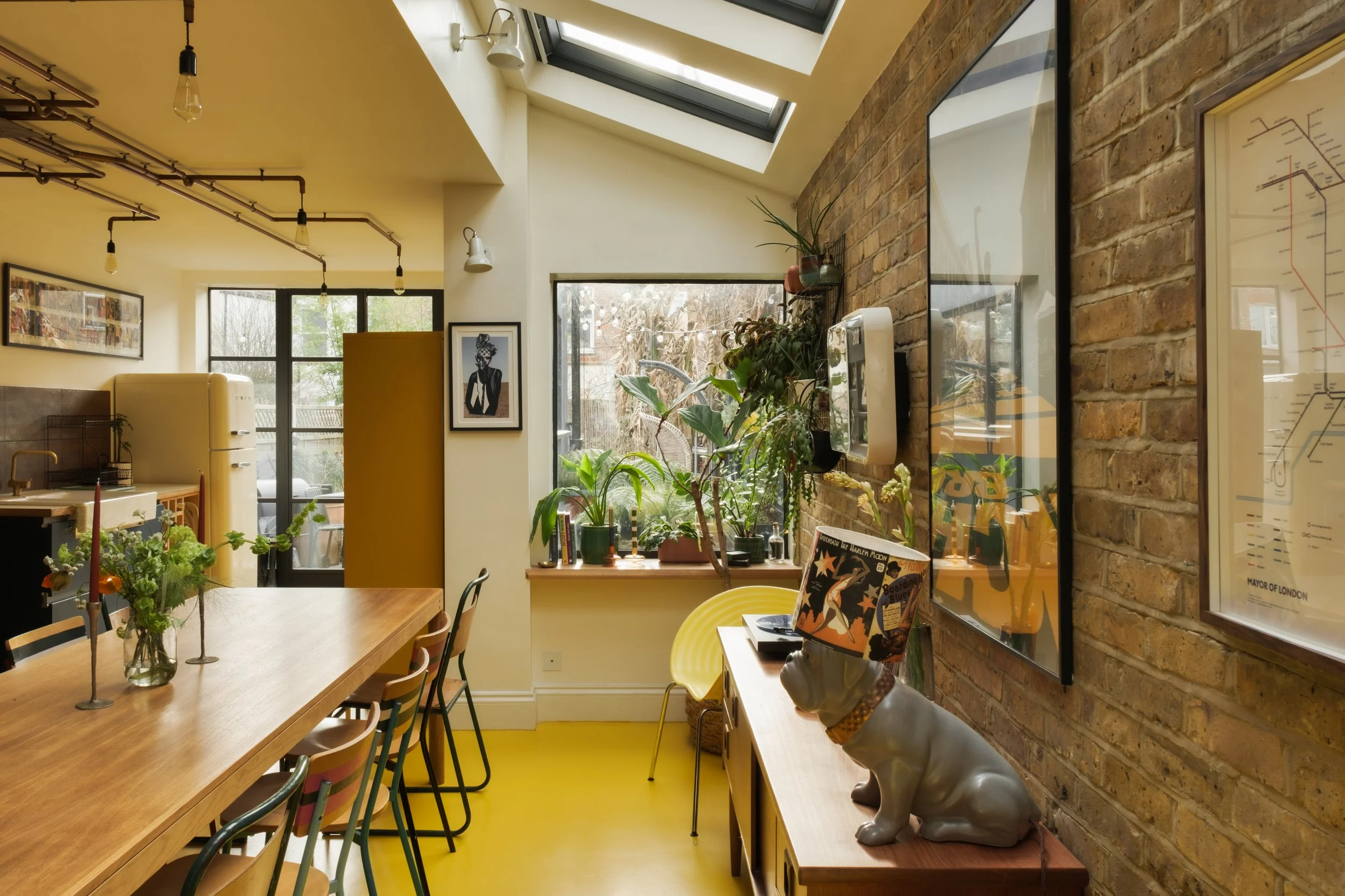 Interior of a cozy, well-lit room with a wooden dining table, various chairs, plants near the window, and decorative artwork on the walls. A porcelain dog statue with a book on its head sits on a sideboard against an exposed brick wall.