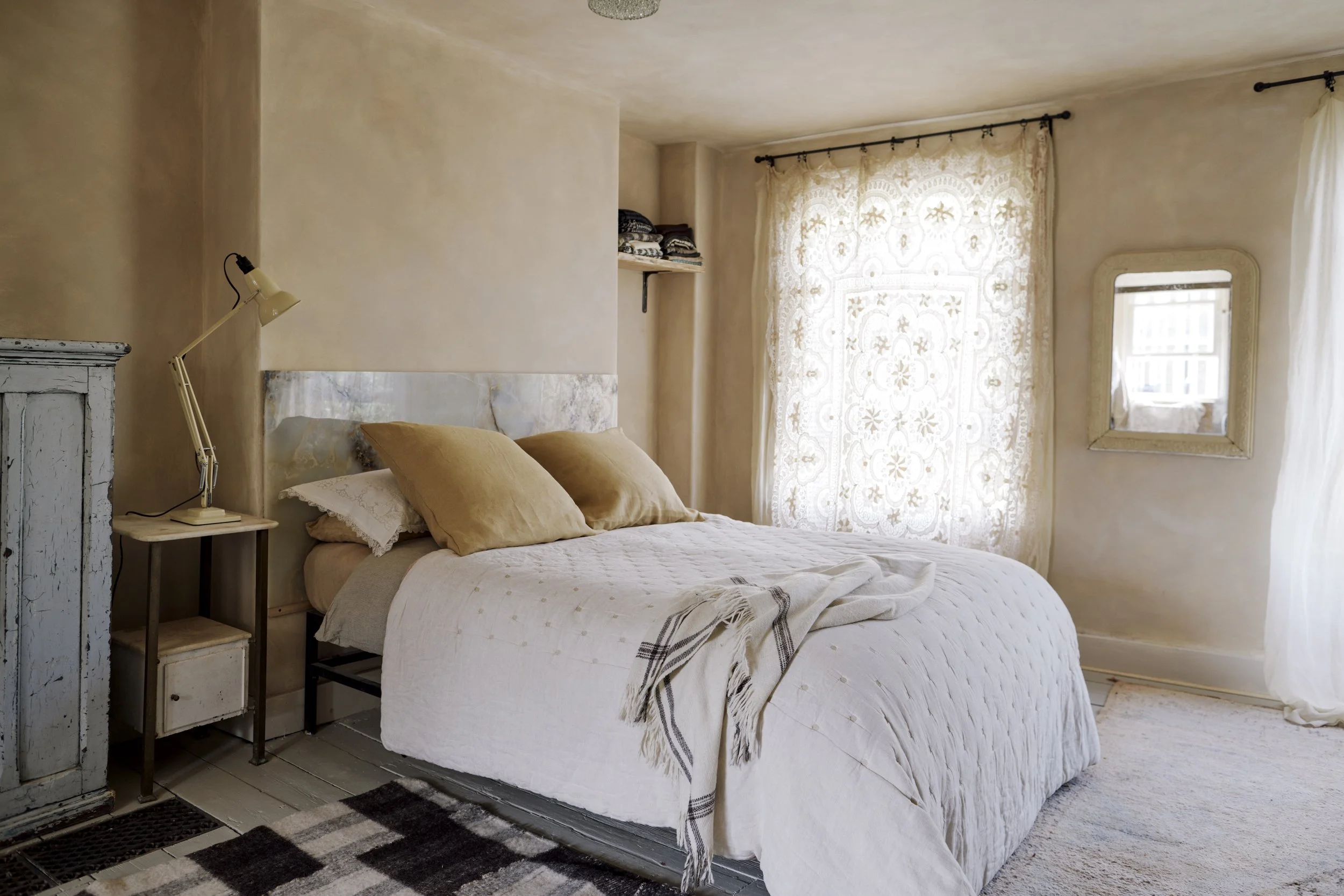 A cozy bedroom with a white bedspread, tan pillows, and a throw blanket, next to a wooden nightstand with a beige lamp, beige curtains with an intricate pattern covering the window, a mirror on the wall, and a striped area rug.