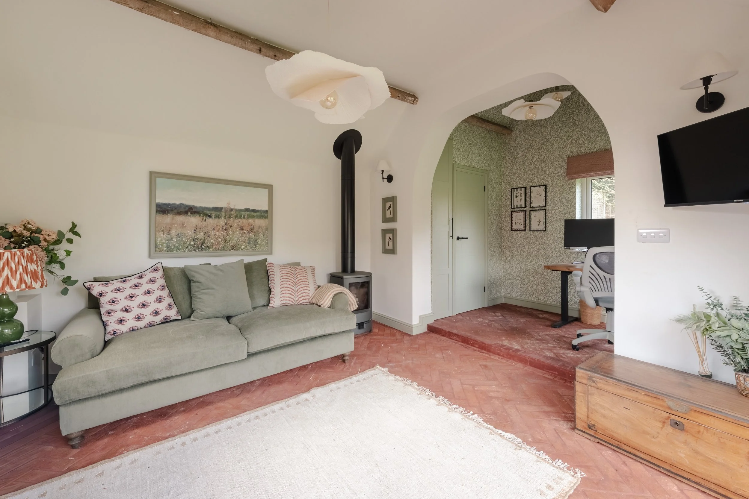 Living room with a light green sofa, patterned pillows, a wooden chest, a black wood stove, and a framed landscape painting on the wall. There is a small side table with a green vase, a lamp, and some plants. The room has white and patterned walls, w
