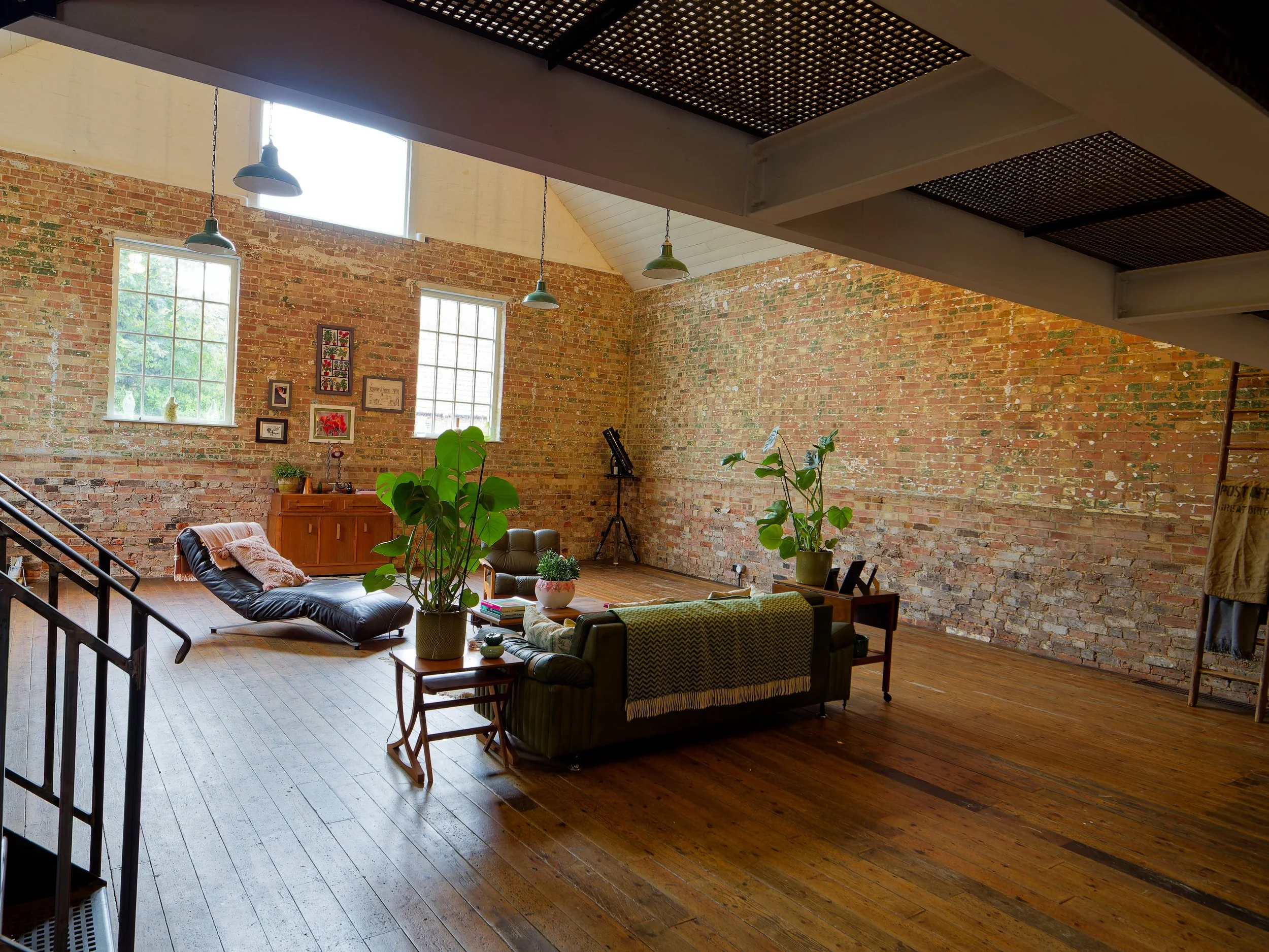 Bright living room with exposed brick walls, large windows, and wooden floors. Furniture includes a green couch, black lounge chair, and potted plants, with framed pictures on the wall.