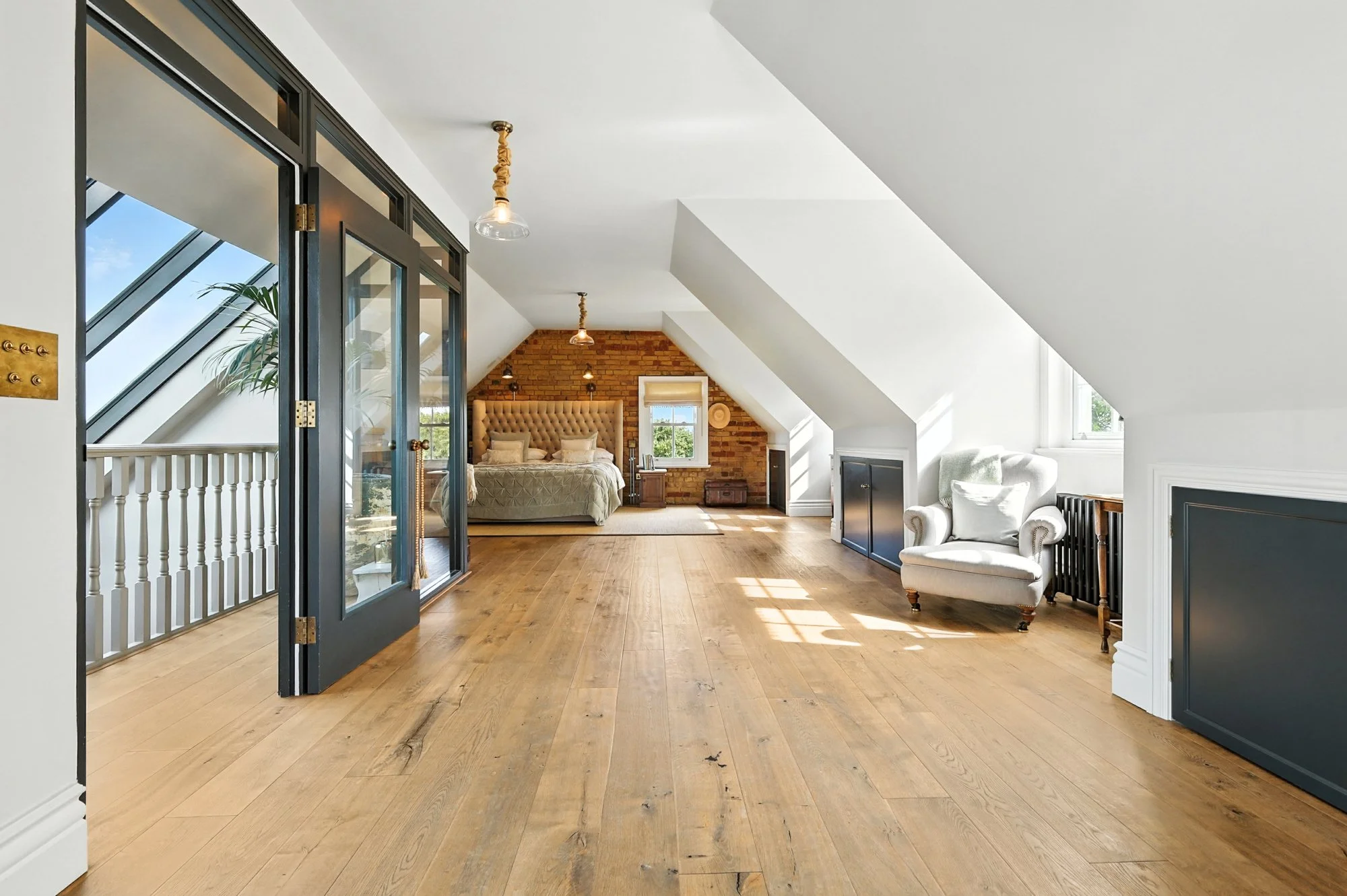 Interior view of an attic or loft space with white walls, sloped ceilings, wooden flooring, a brick accent wall, a large bed with a beige padded headboard, a window with blinds, a white armchair, and modern pendant lights.