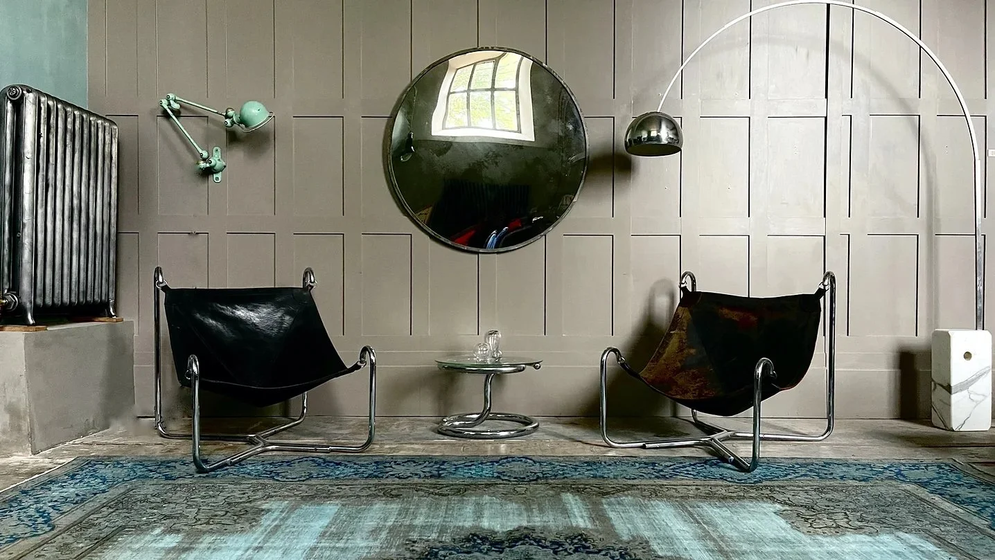 Modern interior space with two black leather chairs, a glass side table, a round mirror, a wall-mounted adjustable lamp, a vintage radiator, and a marble pedestal, with a patterned rug on the floor.