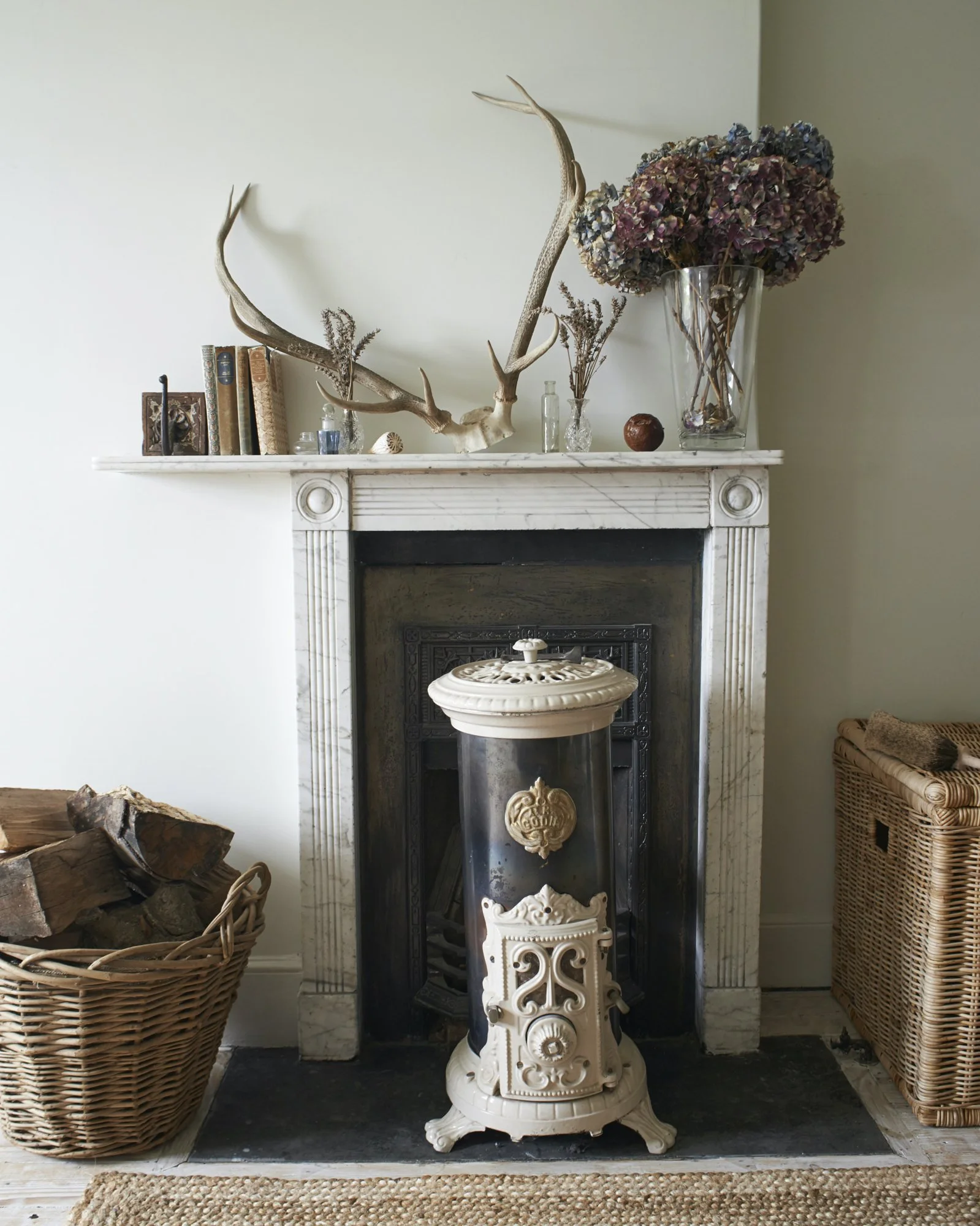 Decorative fireplace with antlers, books, glass bottles, and dried flowers on mantel; vintage heater in front; basket of firewood to the side; wicker furniture on right.