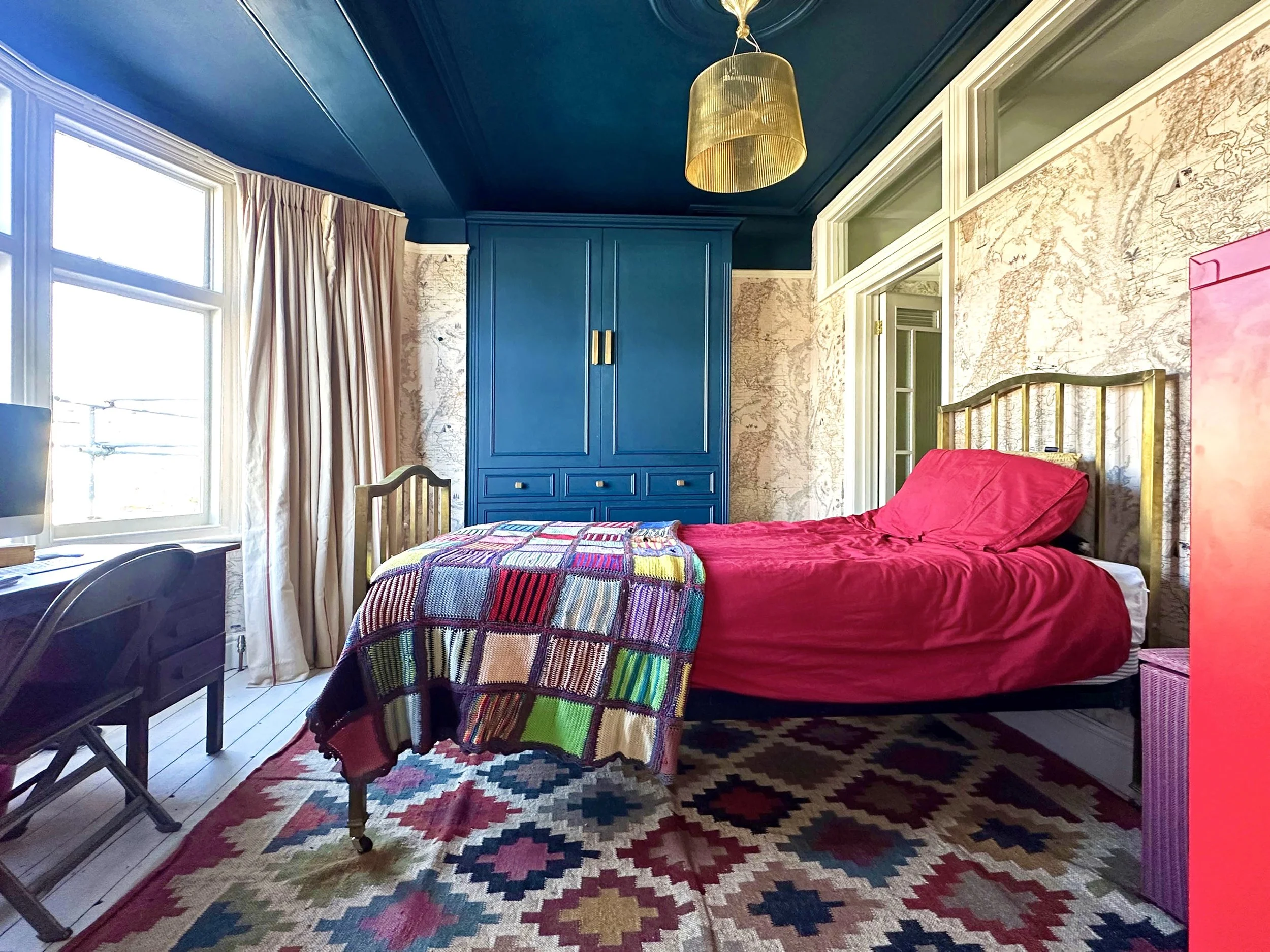 A bedroom with blue ceiling and walls, a bed with red bedding, a colorful blanket, a large patterned rug, a blue wardrobe, a window with curtains, and a desk with a computer.