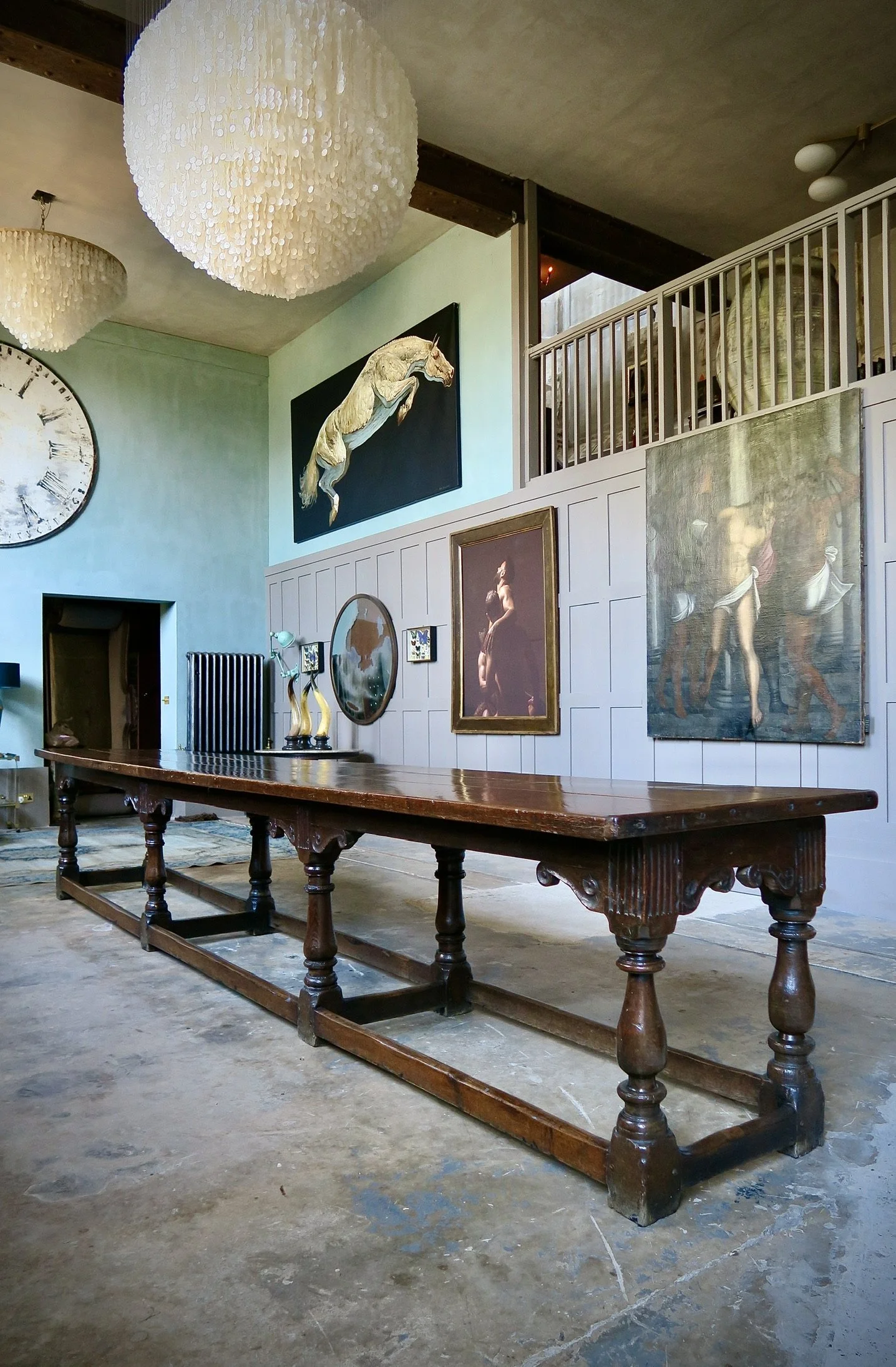 An interior room with hanging lights, a large wooden table, wall art including a clock and paintings, and a staircase or railing.