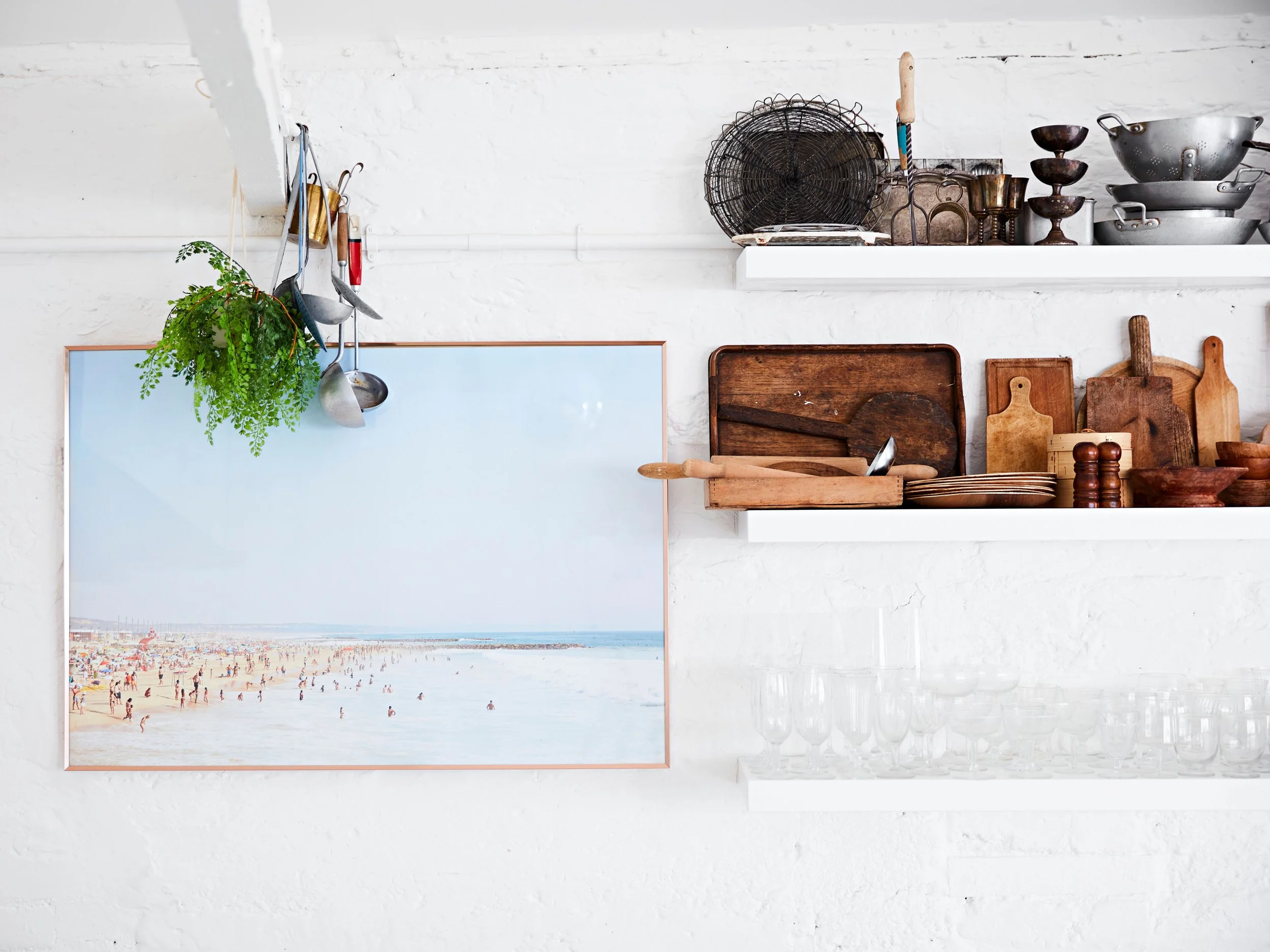 Interior of a kitchen with white brick wall, open shelves holding wooden cutting boards, metal bowls, and vintage kitchen tools, and a large framed beach scene photo.
