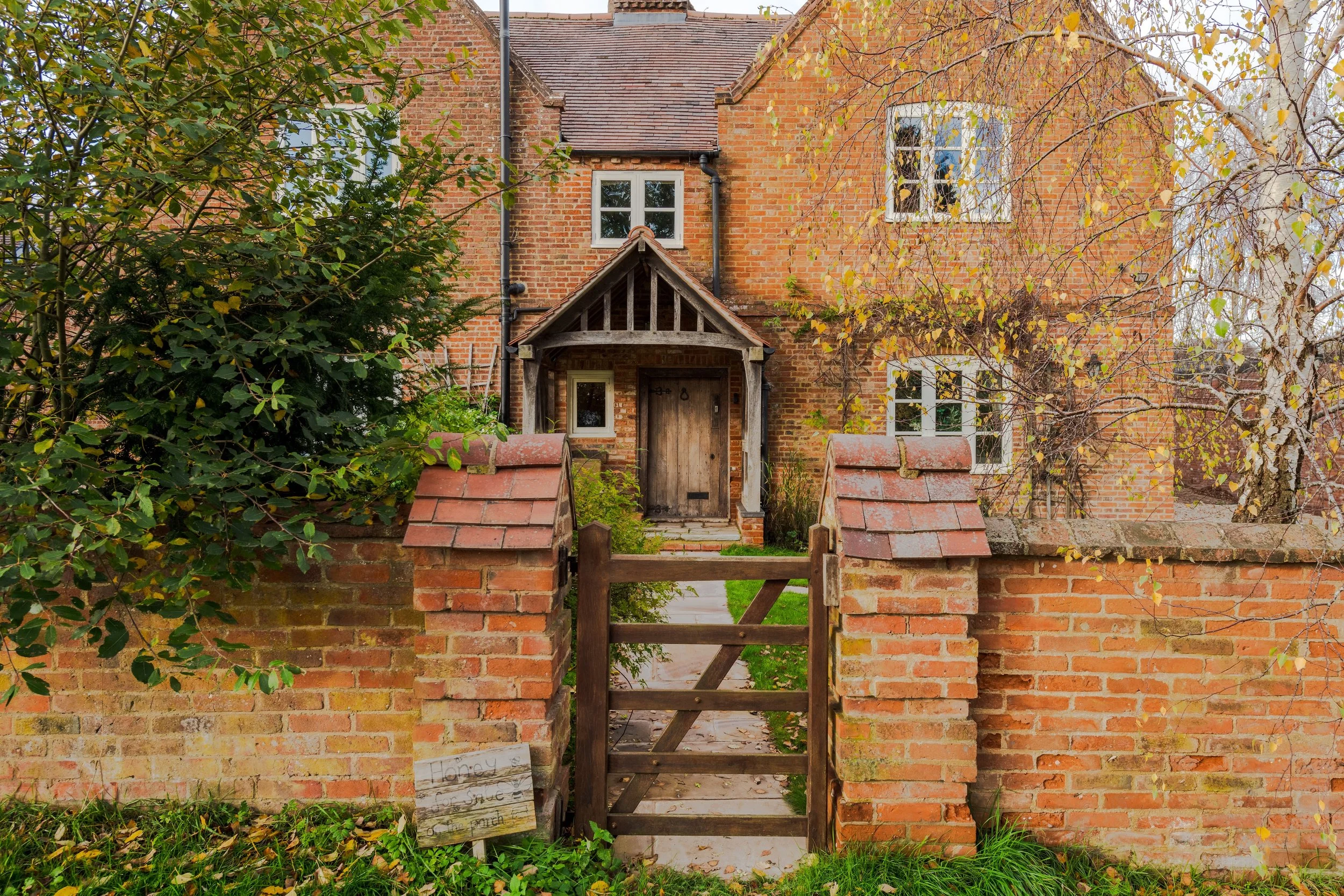 Brick house with front gate and brick wall, surrounded by trees and plants, autumn leaves on the ground.
