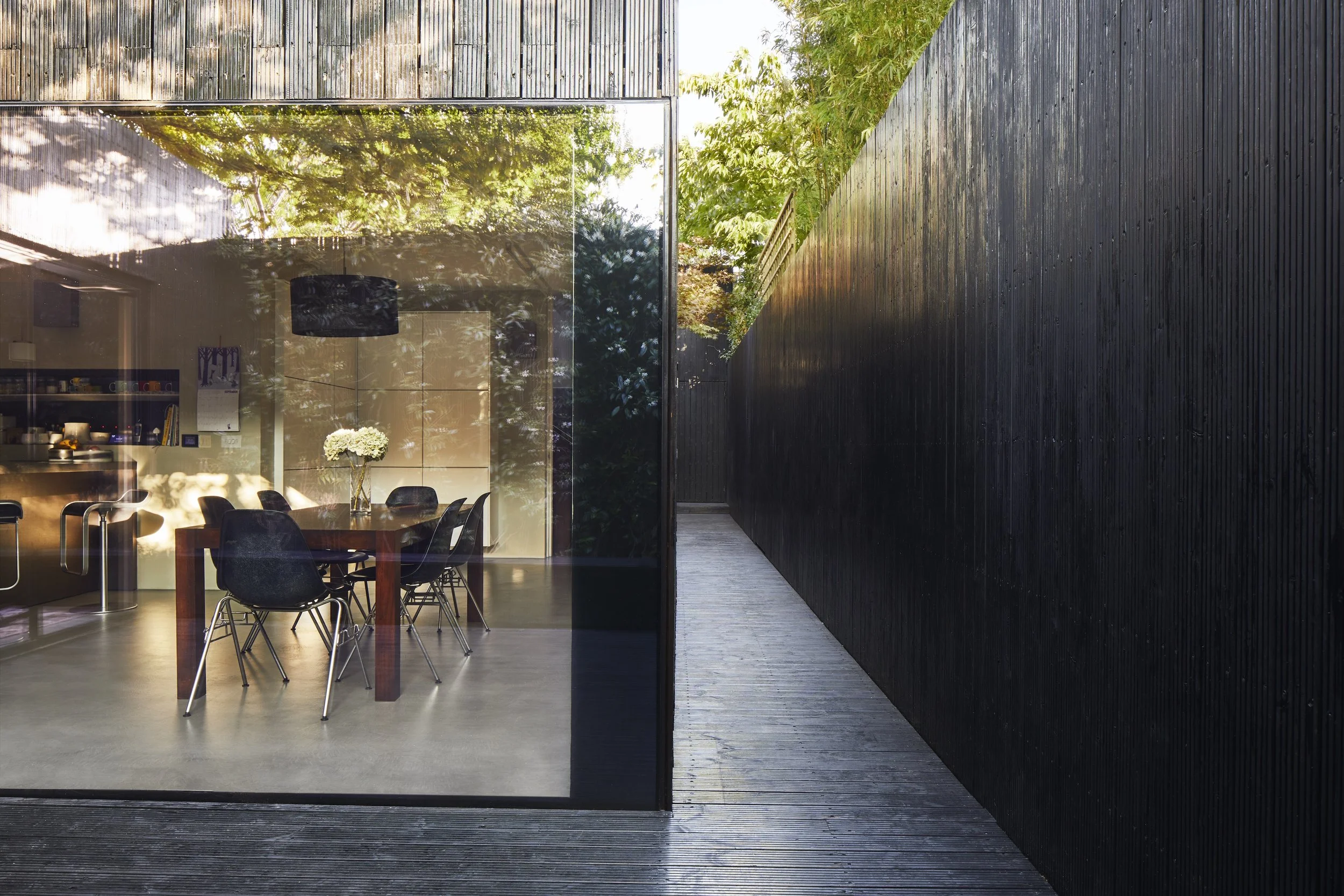 Modern dining room visible through a large glass window, with a wooden table, six black chairs, and a vase of white flowers, adjacent to a black wooden exterior wall and a wooden walkway surrounded by greenery.