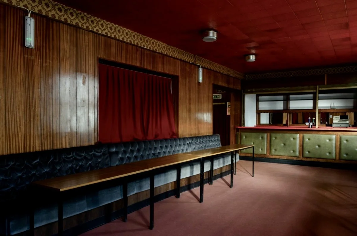 Empty bar or lounge area with wood-paneled walls, a long black cushioned bench, small tables, a red curtain, and a green-paneled bar in the background.