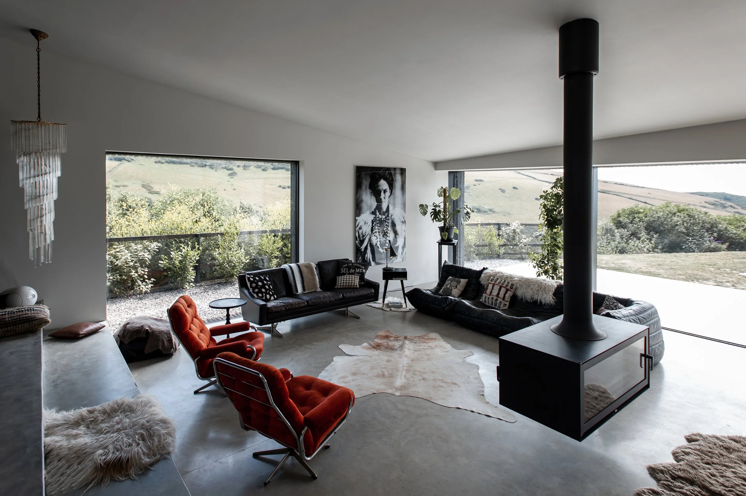 Modern living room with large windows showcasing a scenic outdoor view, featuring black and red chairs, black sofas, a cowhide rug, and a potted plant, with a black fireplace and artwork on the wall.
