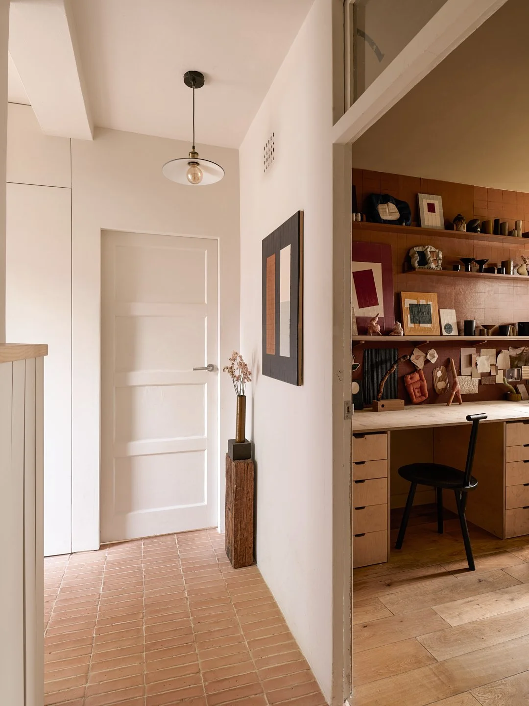 Interior view of a home with a hallway and a workspace. The hallway has a white door, a ceiling light fixture, and a tall vase with dried flowers. The workspace features wooden shelves with various decorative items, framed artwork, and a black chair.