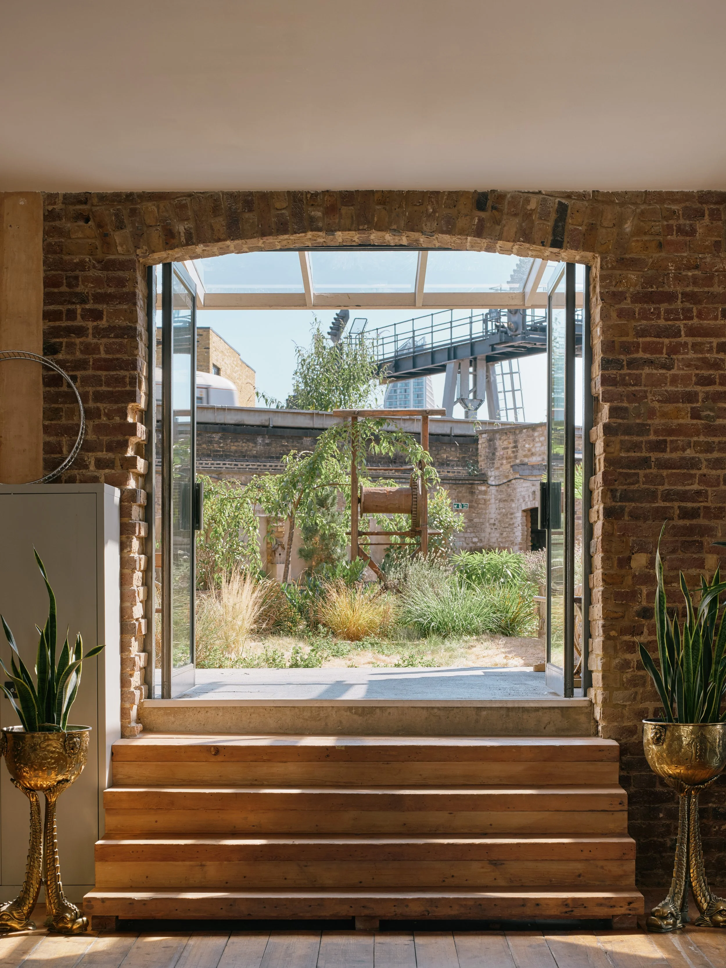 Looking out from inside a brick-walled room through an open glass door at a garden with plants, trees, and a playground slide, with a building and a crane in the background.
