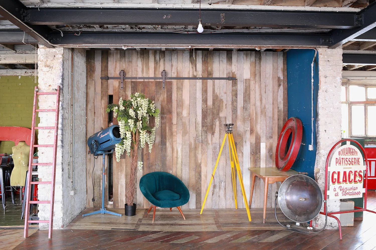 Interior of a rustic room with wooden wall, vintage furniture including a teal armchair, a small wooden table, and a standing light; a yellow tripod and a large reflector on the floor; decorative plants; a pink ladder and a sign that reads in French 